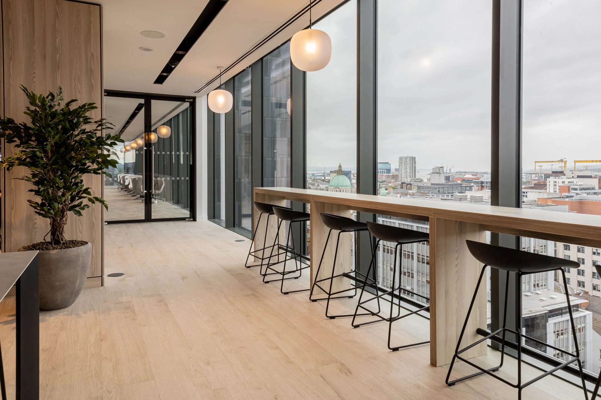 Real estate photography of modern office with floor-to-ceiling windows, black barstools along a wooden counter, and city skyline view on a cloudy day.