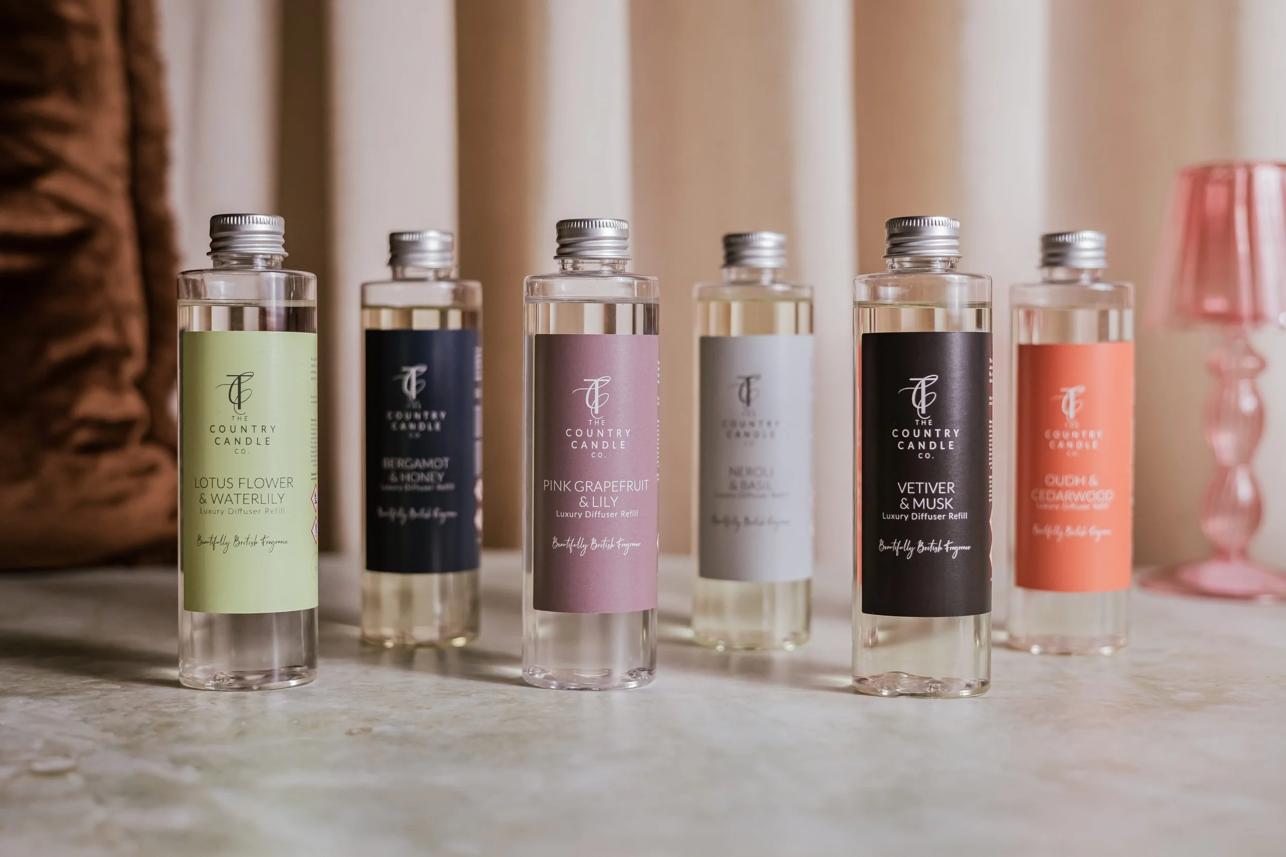 Six bottles of luxury reed diffusers with different scents placed on a table, including Lotus Flower & Waterlily, Pink Grapefruit & Lily, Vetiver & Musk, and other scents, with a pink decorative candle holder in the background.