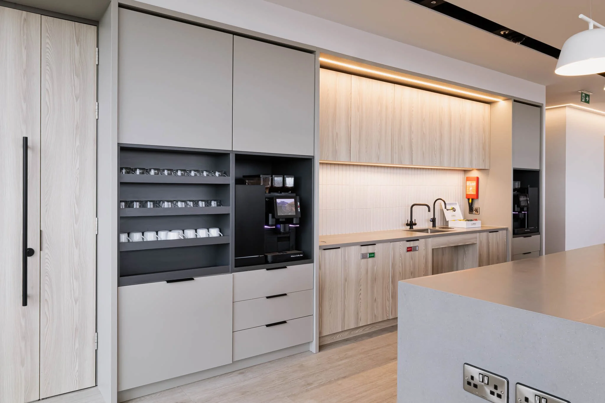 A modern coffee station with beige and light wood cabinetry, a coffee maker, cups and glasses, and a double sink with black faucets captured for building photography UK