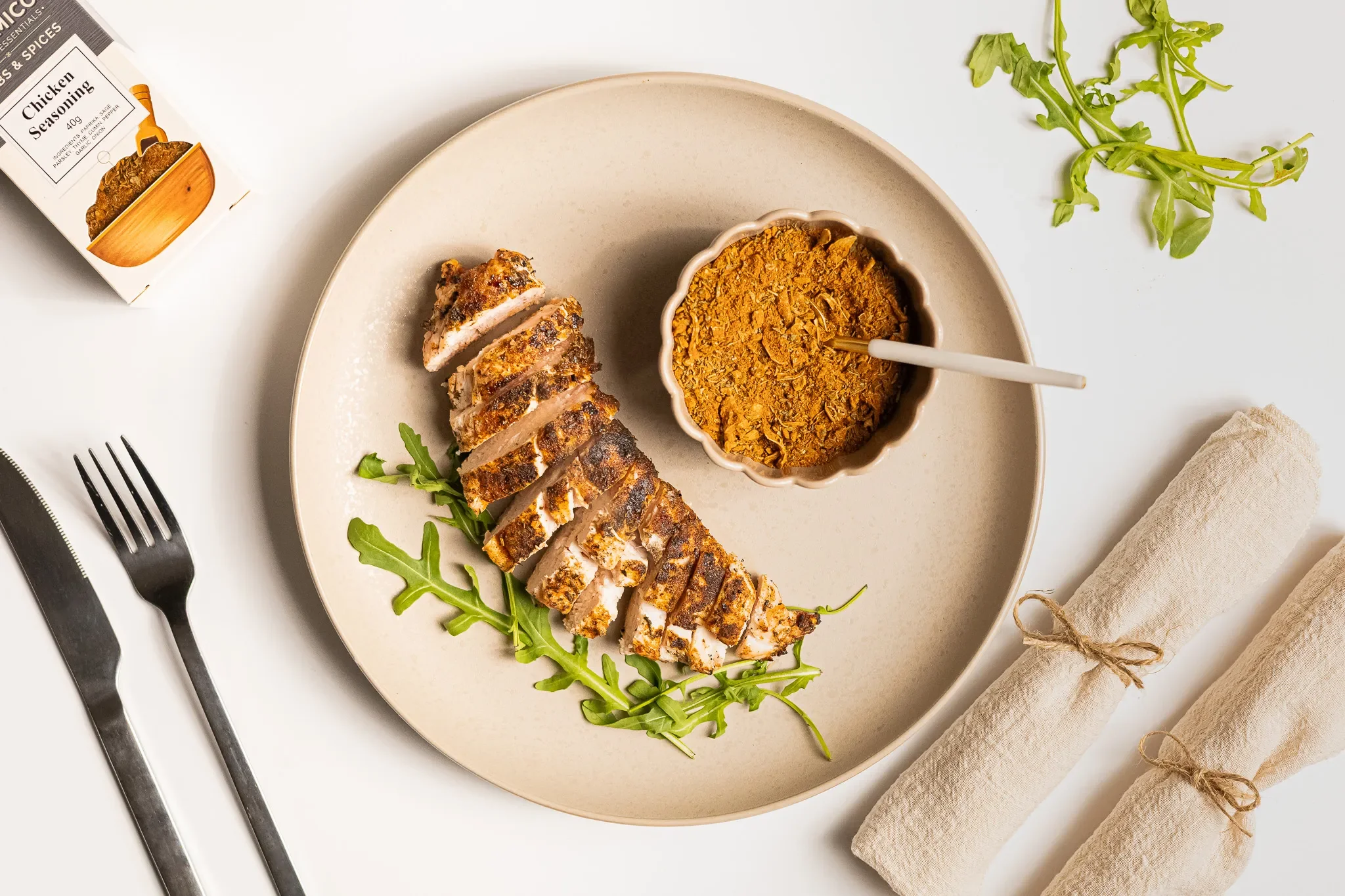 Sliced cooked chicken breast on a beige plate with arugula, served with a bowl of curry on the side, on a white table. Silver fork and knife, two rolled cloth napkins, and a spice jar are also on the table.