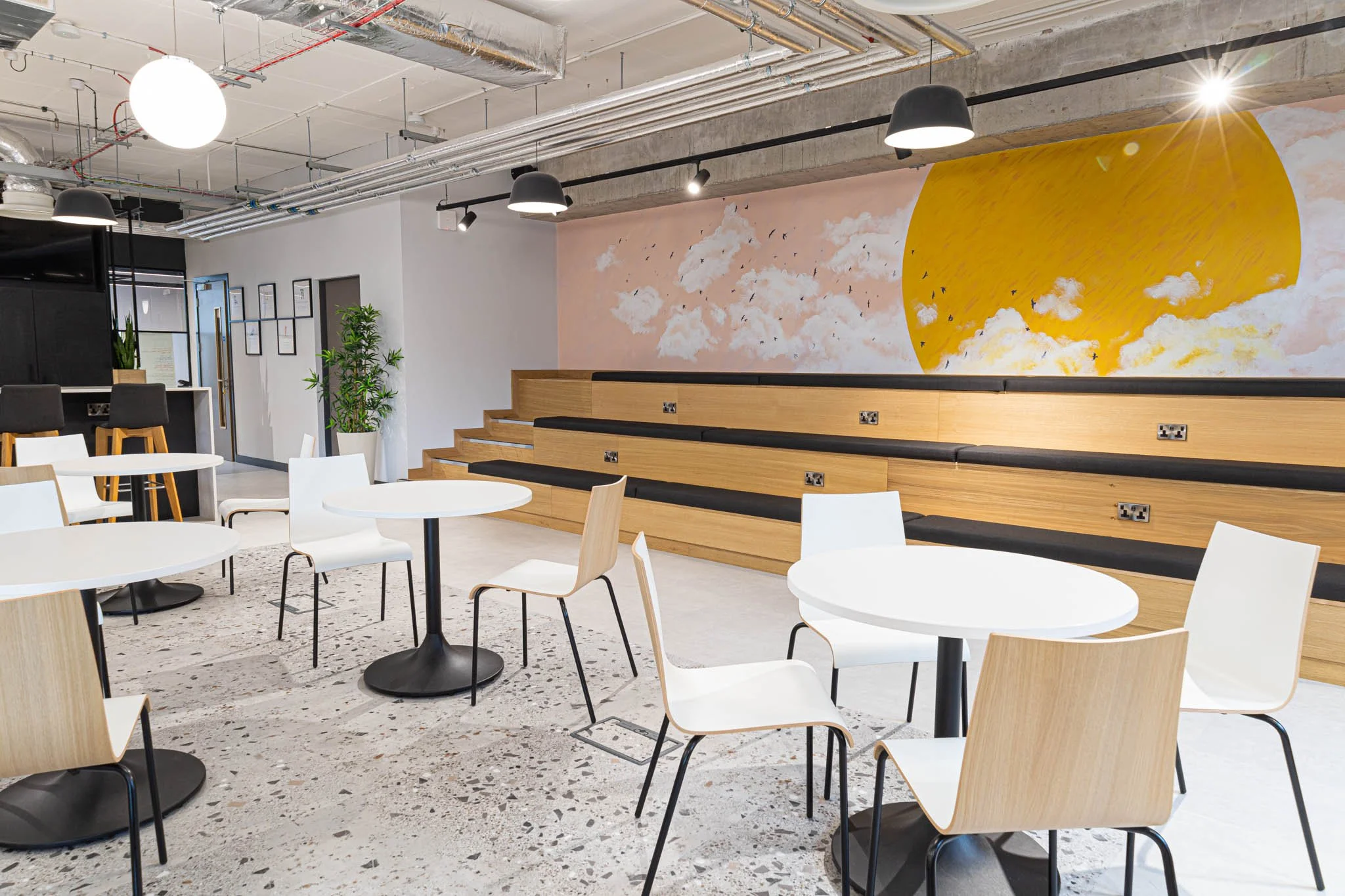 Modern coffee shop interior photography with white round tables, light wooden chairs, and tiered wooden seating with black cushions, featuring a large mural of a partly cloudy sky and a sun on the wall.