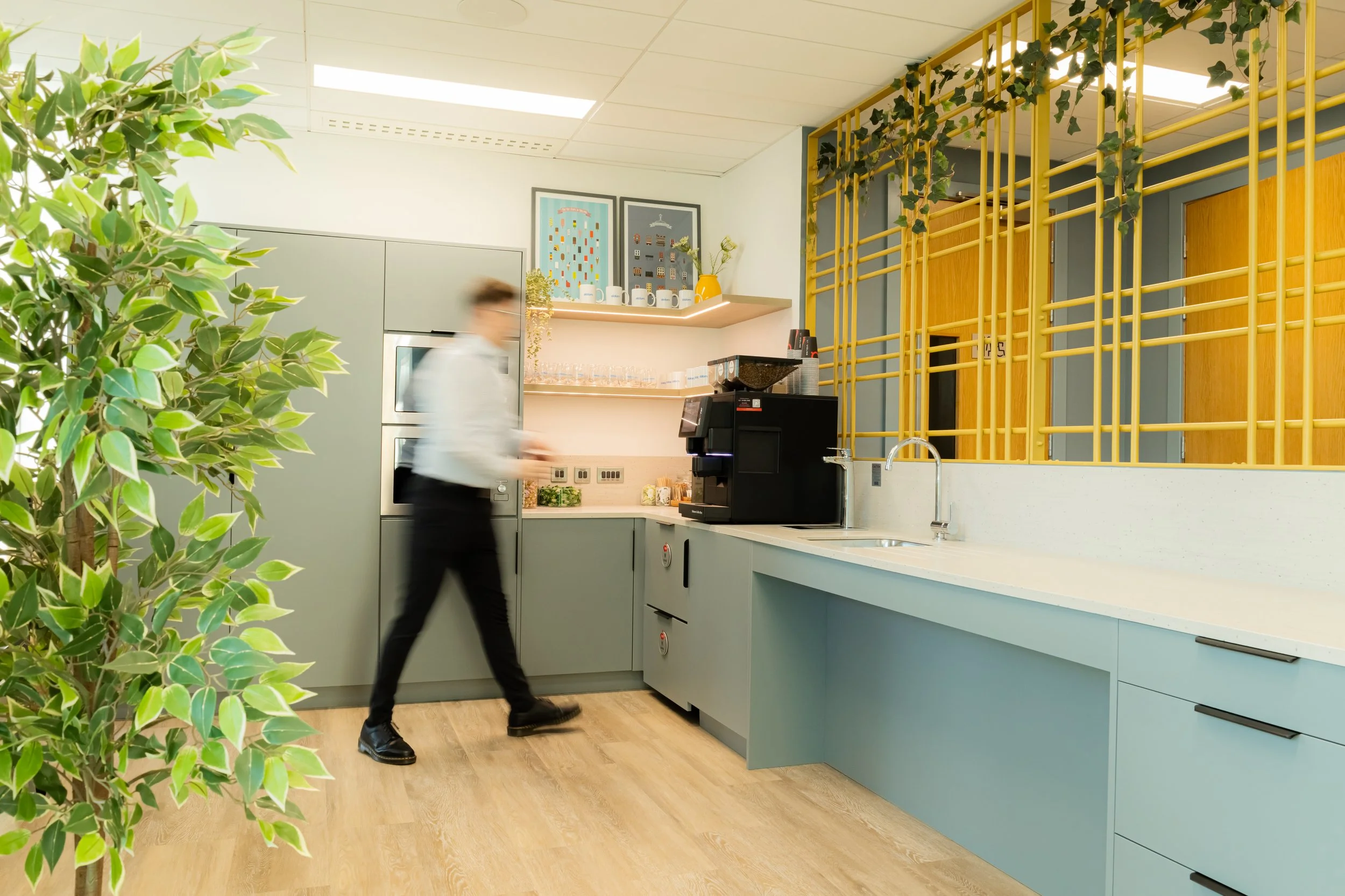 Property photography of modern office kitchenette with a person walking past, plants, grey cabinets, wooden flooring, decorative pictures on the wall, and a coffee machine on the counter.