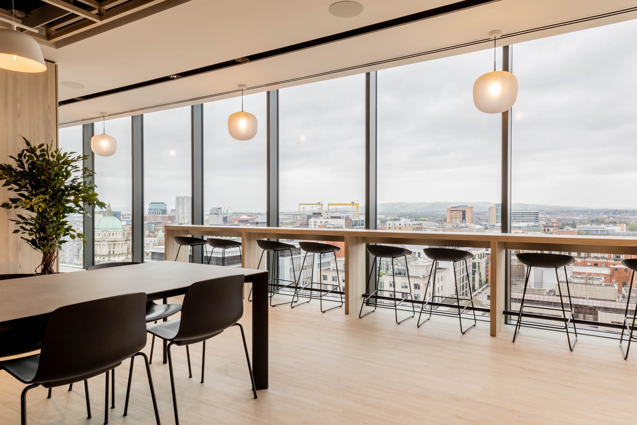 Modern office space with large windows overlooking a city skyline, black chairs, a long wooden table, hanging pendant lights, and a potted plant.