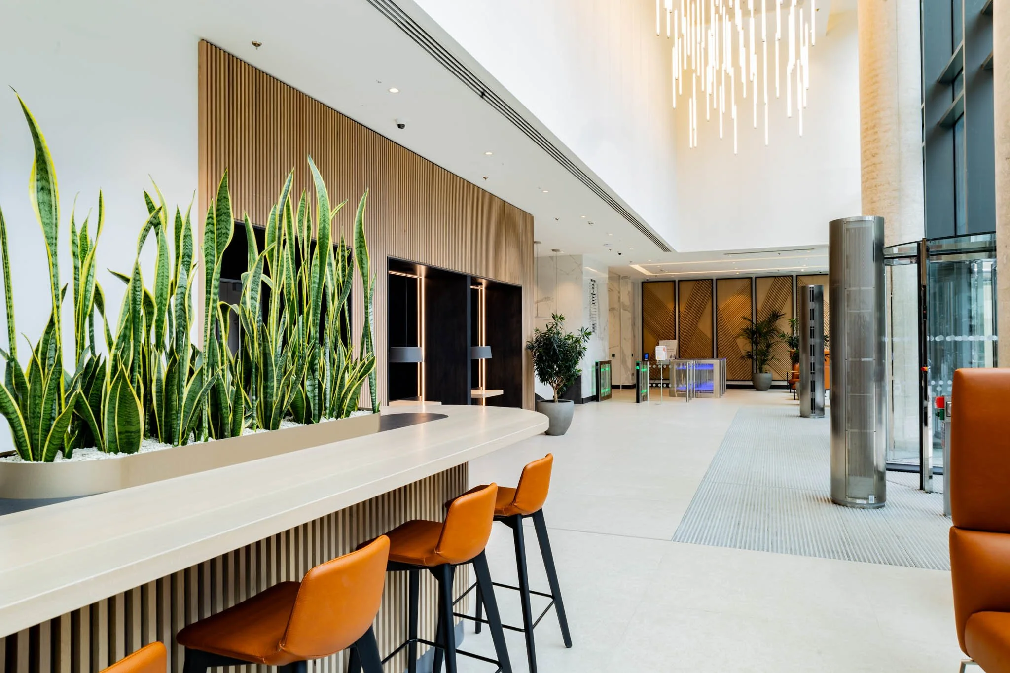Modern hotel lobby with large plant, orange chairs, wood and marble decor, and a high ceiling with a hanging light fixture captured by a hotel interior photography UK service