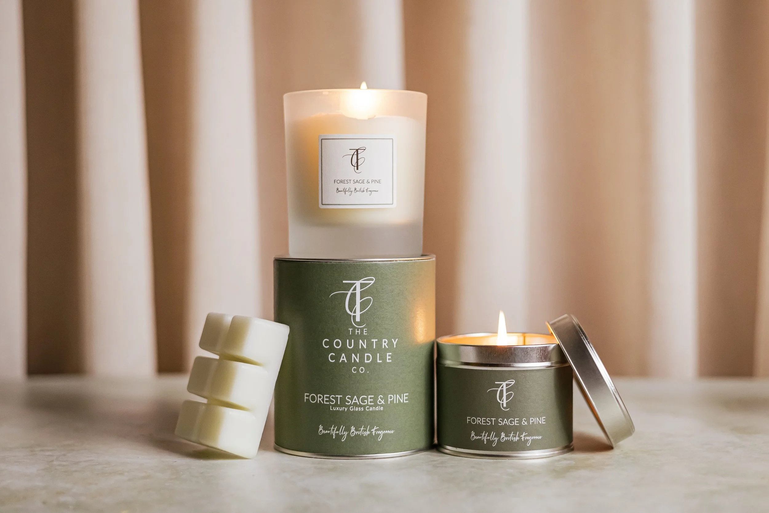 A set of candles and wax melts on a table. The candles have labels reading "Forest Sage & Pine" and "The Country Candle Co." with one candle lit. There are white wax melts stacked on the left. The background is blurred with beige curtains.