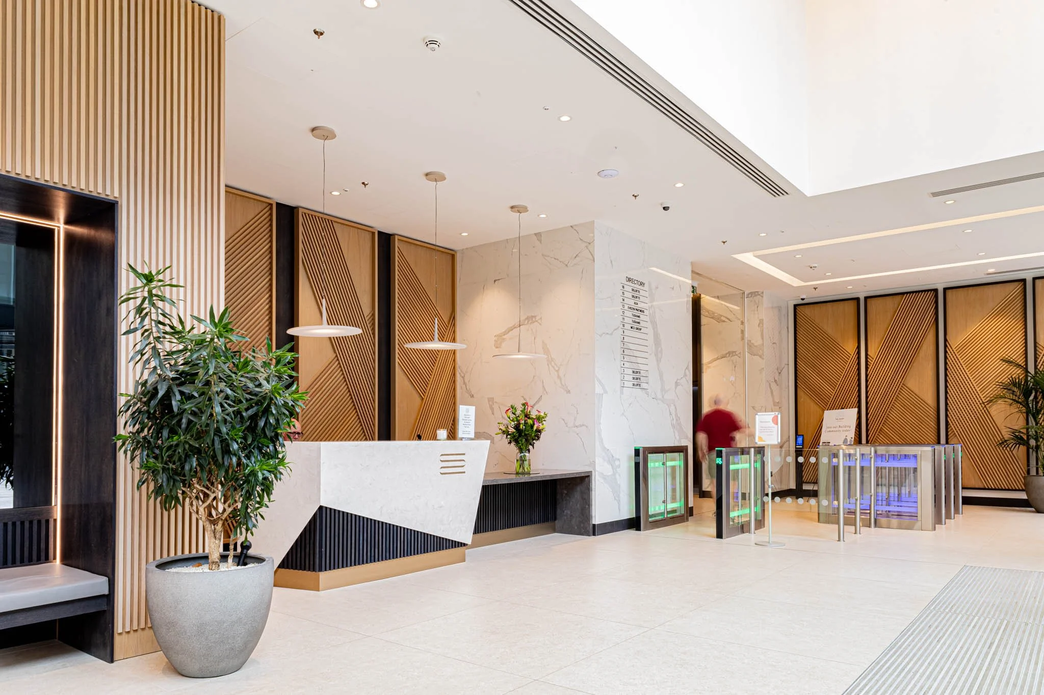 Hotel interior photography of lobby with modern decor, reception desk, potted plants, flower arrangement, and automatic turnstiles at entrance.