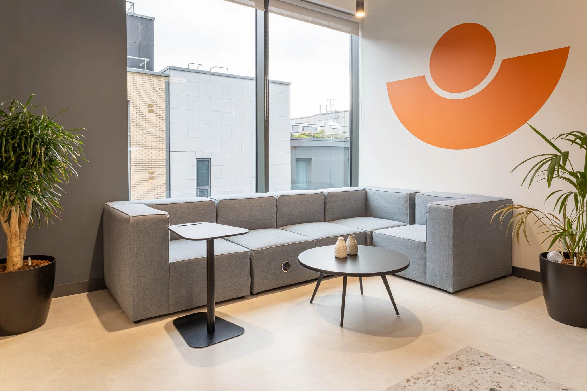 Modern office lounge area with a grey sectional sofa, two black coffee tables, large window with city view, and two potted plants, one on each side of the sofa, with a large orange abstract wall art, captured by an interior photographer UK