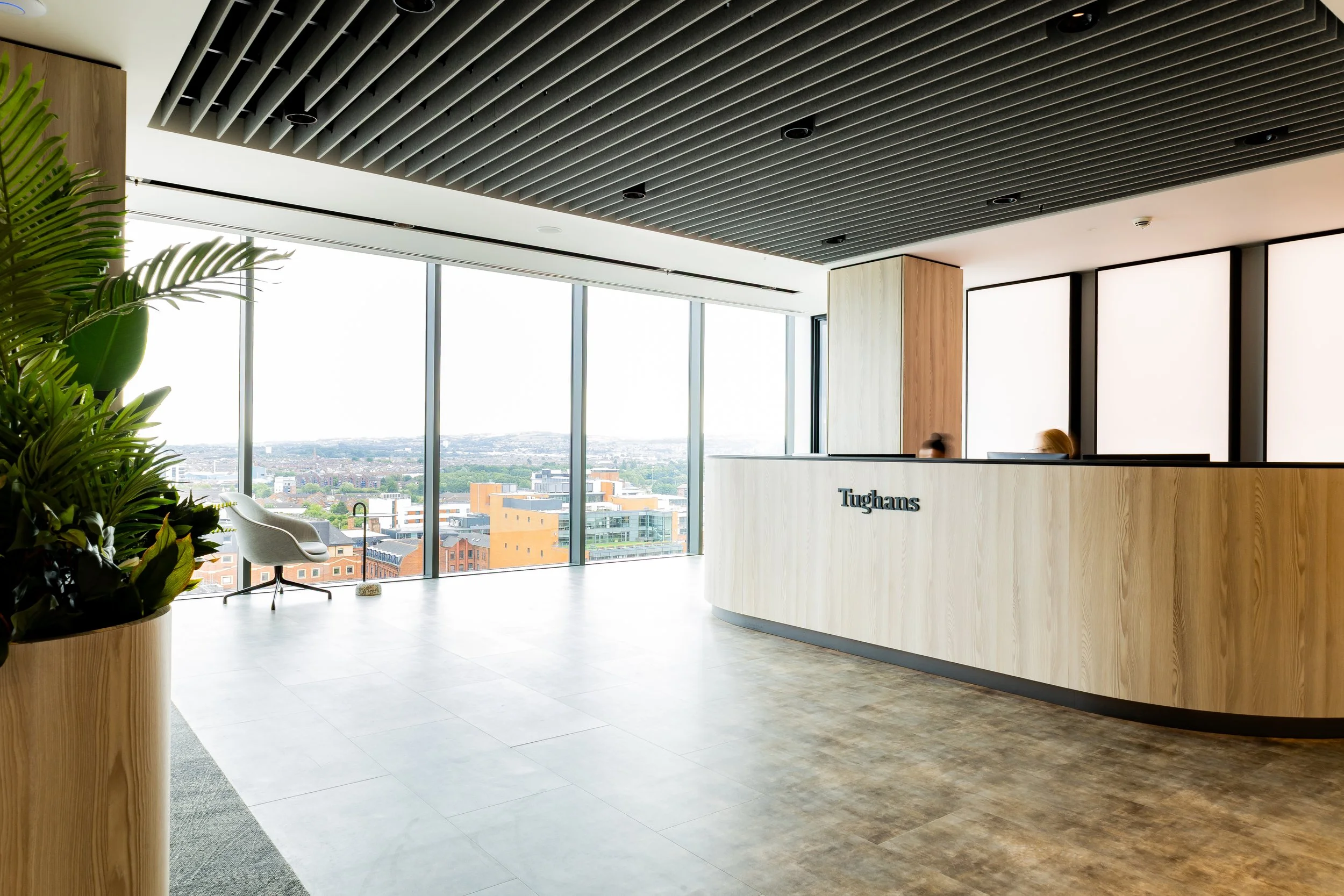 Interior photography uk - Modern office interior with a curved wooden reception desk displaying the name 'Tughans', large windows with city skyline view, and indoor plants in belfast.