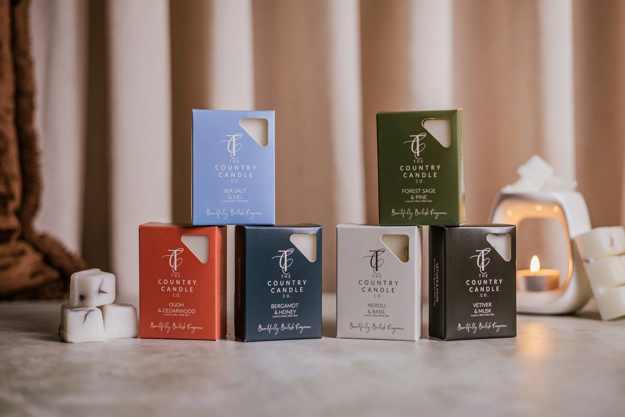 Six boxes of luxury wax melts from The Country Candle Co, in various scents arranged on a table with a black and white candle in the background, and three white wax melts stacked in the foreground.