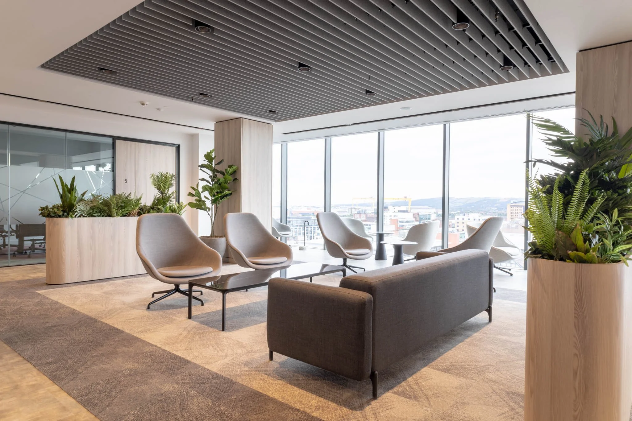 Interior photography of modern office lounge area with beige chairs, a dark gray sofa, and large window with city views, decorated with green plants in belfast.