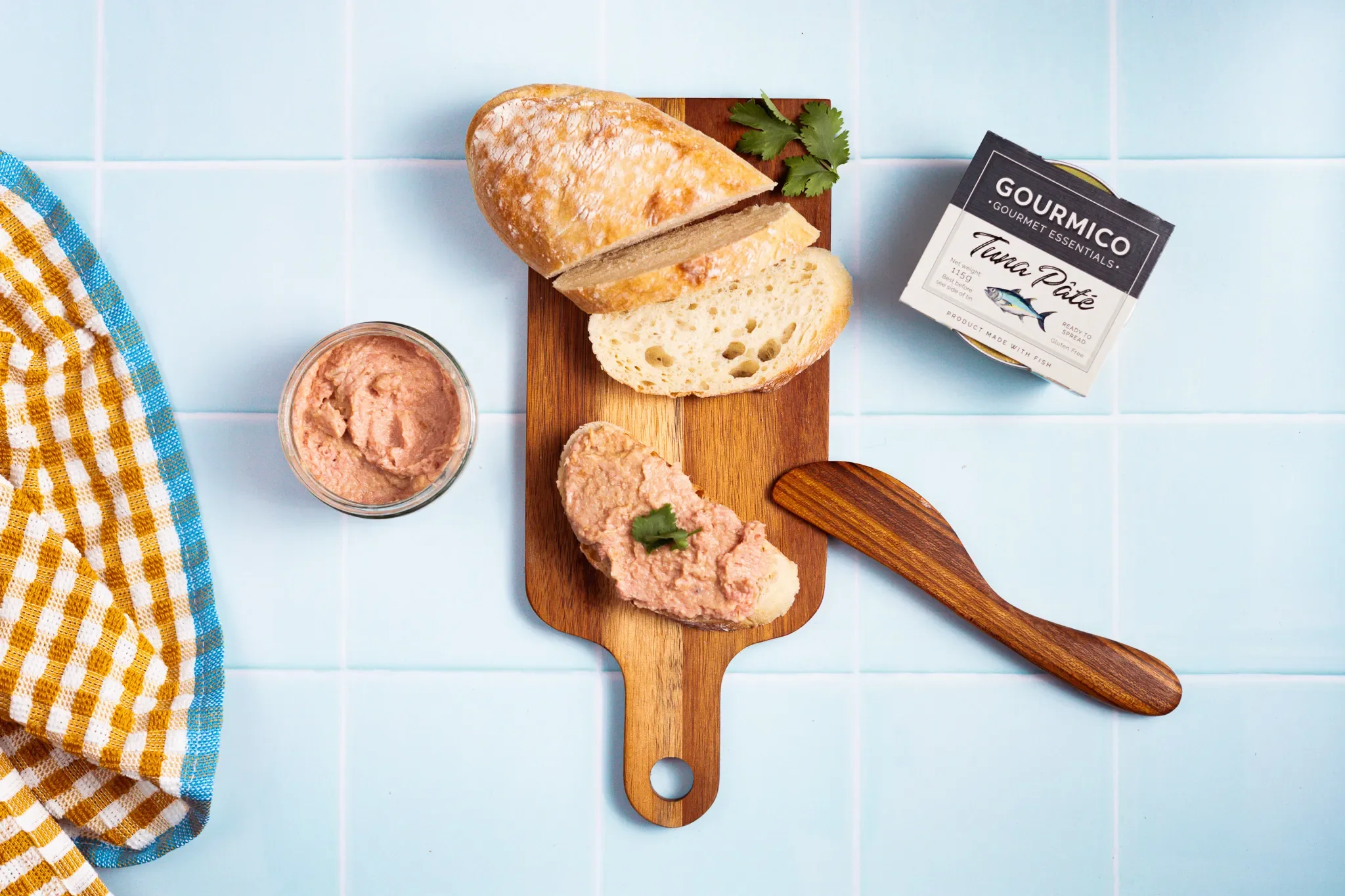 A wooden cutting board holding a loaf of bread, a slice of bread, and a generous spread of fish pâté, with parsley for garnish, on a light blue tiled surface, with a jar of pâté, a box of gourmet fish spread, and a colorful checkered cloth nearby.