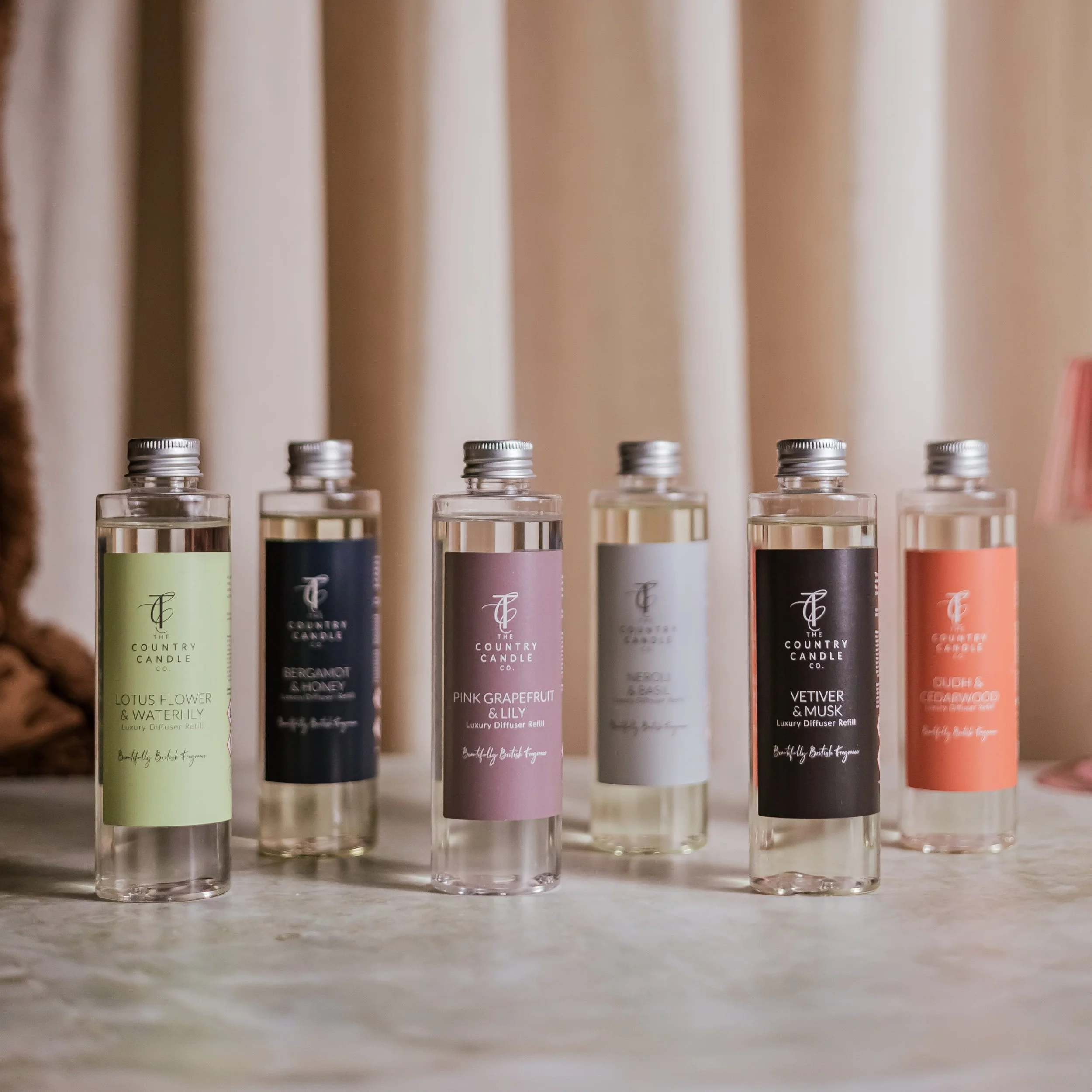 Six bottles of luxury diffuser refills labeled with various scents, arranged in a row on a table with light-colored curtains in the background.