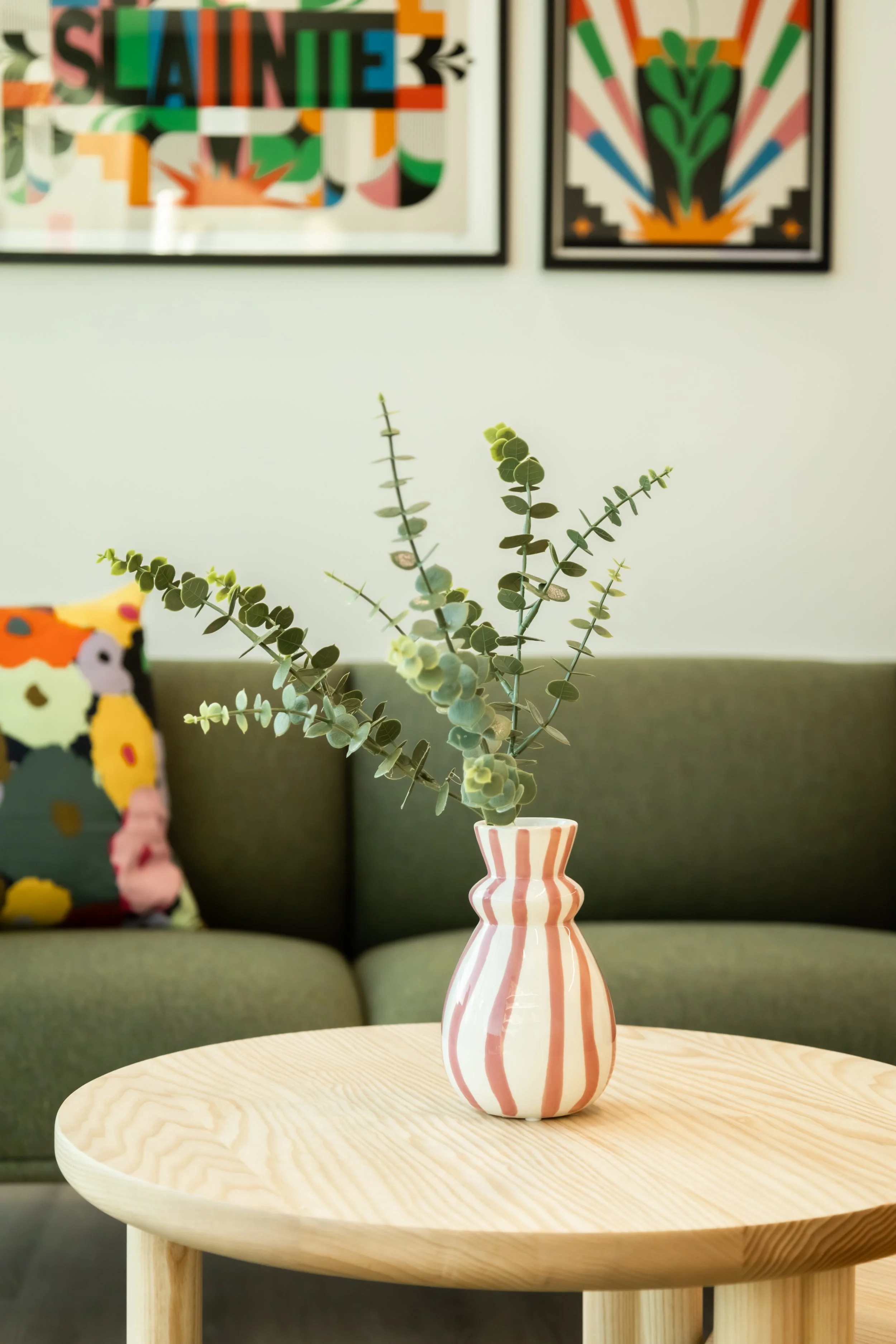 A green leafy plant in a striped white and pink vase sitting on a round wooden table in a living room with a green couch and colorful framed artwork on the wall.