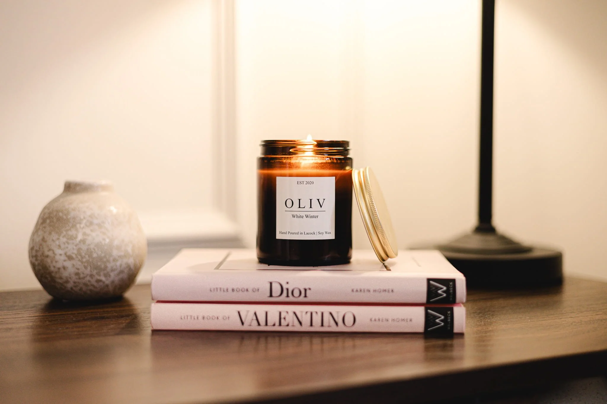 A lit brown candle labeled 'OLIV White Winter' placed on top of two stacked books titled 'Little Book of Dior' and 'Little Book of Valentino' on a wooden surface, with a small white textured vase on the left and a black lamp on the right.