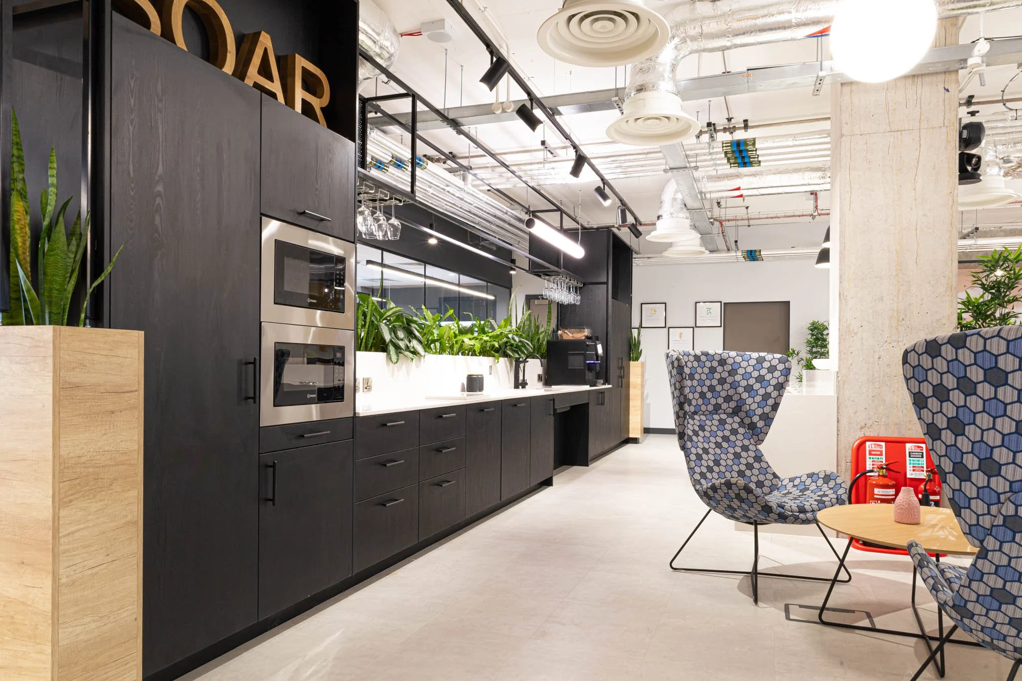 Modern office kitchen area with black cabinets, built-in microwave and oven, green plants, and chairs with patterned upholstery around a small table, fire extinguisher nearby.