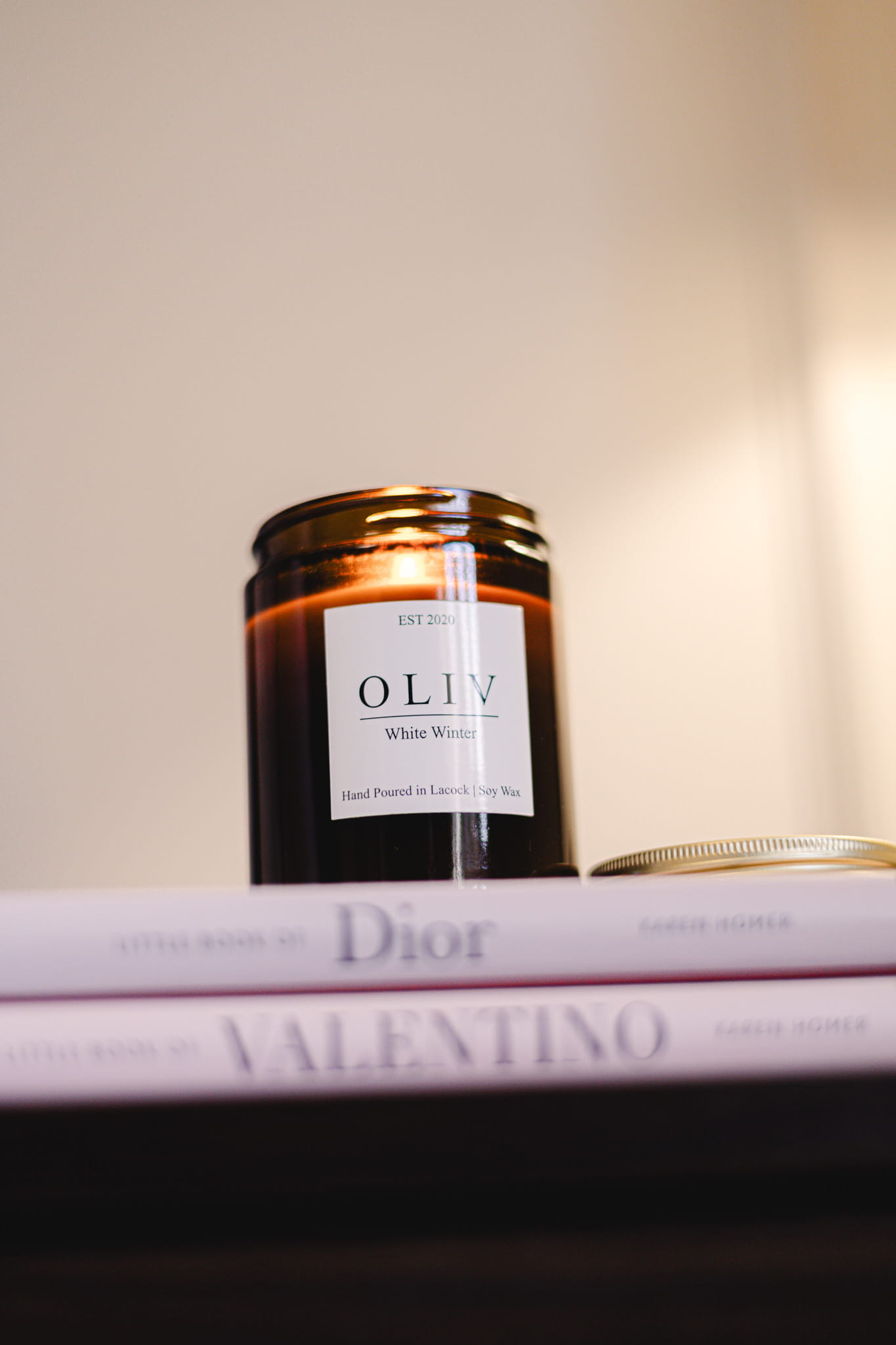 A brown glass candle jar labeled "OLIV White Winter" on top of two fashion magazines, 'Dior' and 'Valentino', with neutral-colored walls in the background.