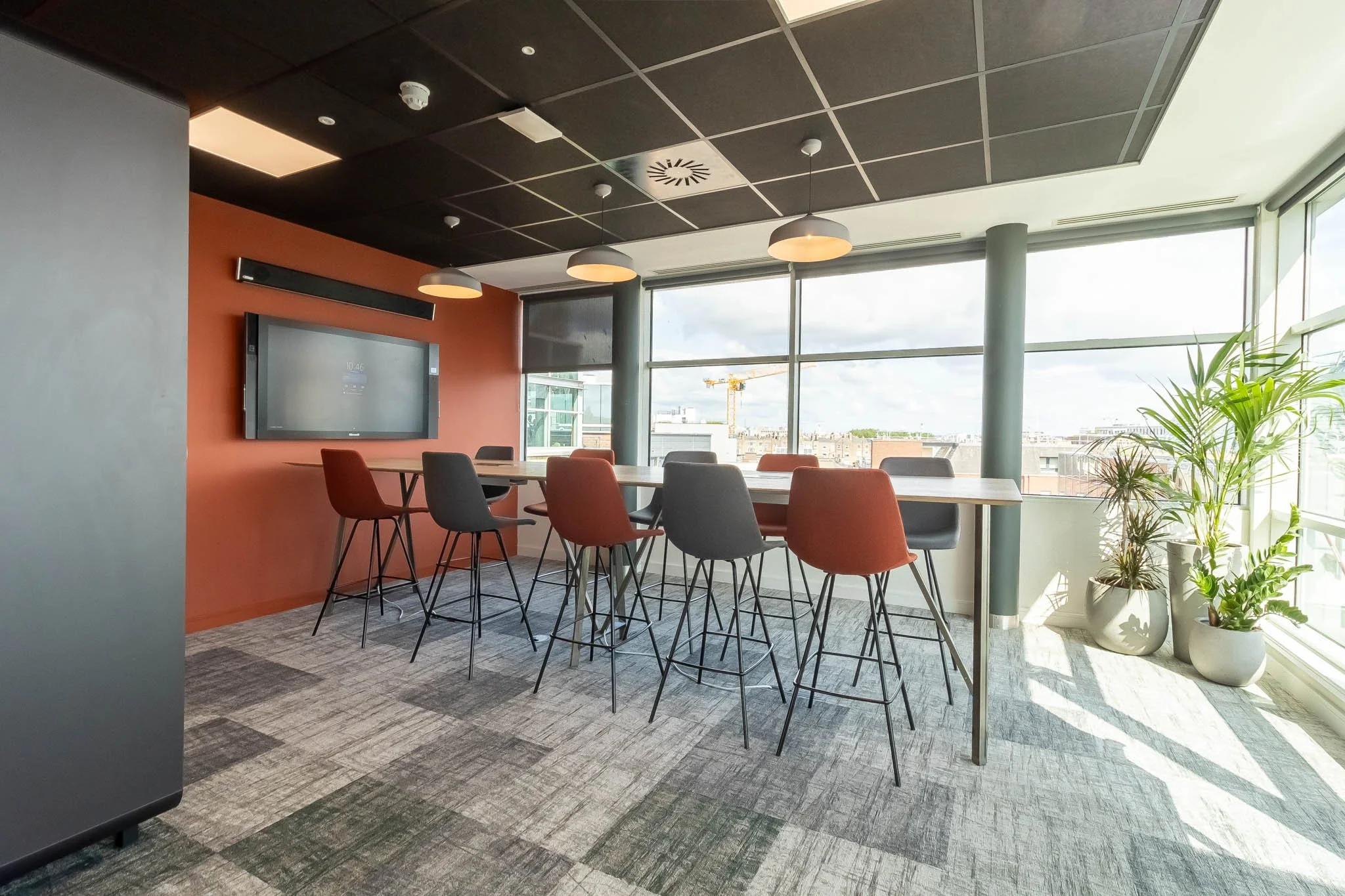 Modern meeting room with high table, red and gray chairs, floor-to-ceiling windows, and potted plants outside sunlight.