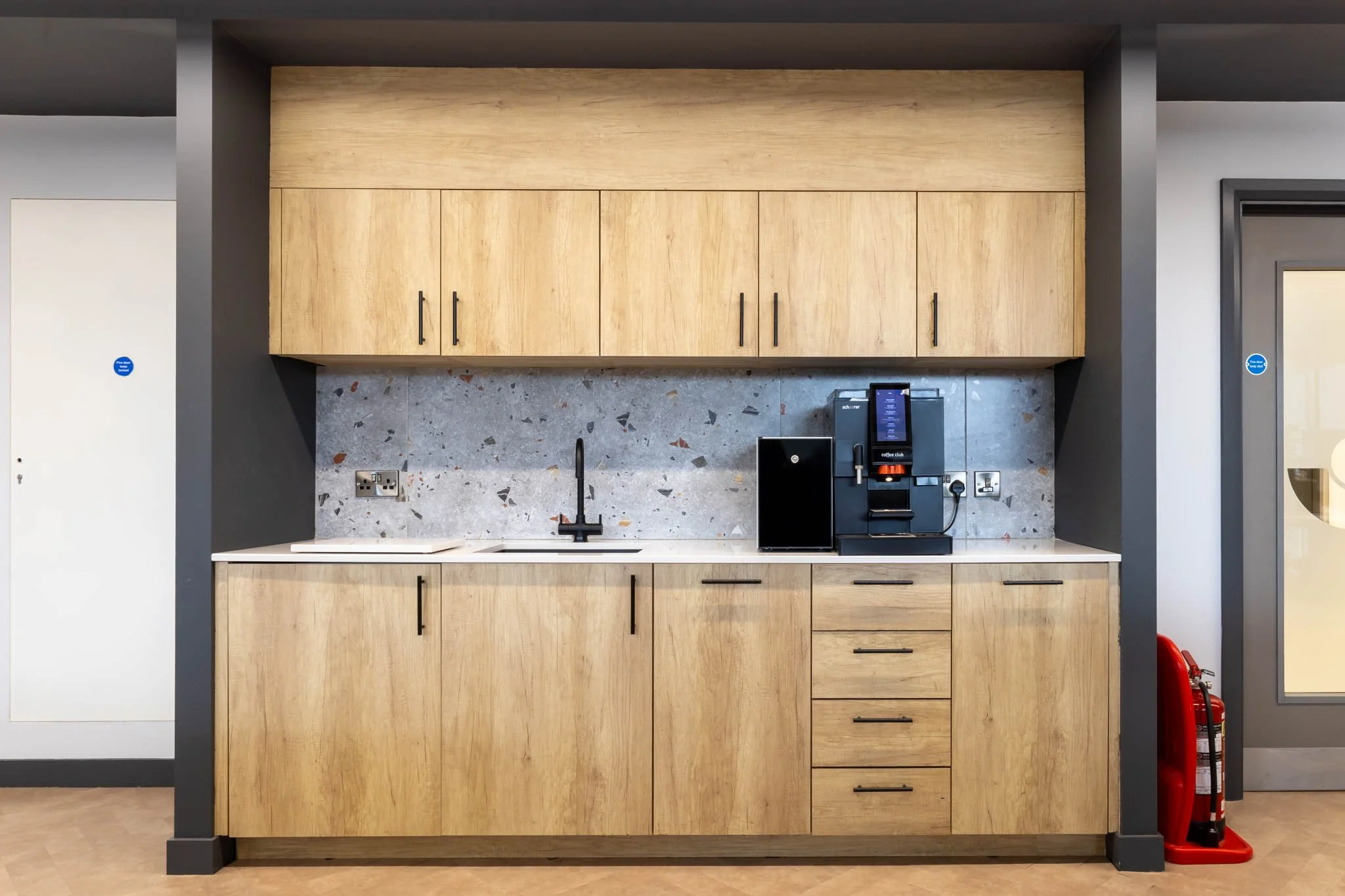 Interior photography of kitchen area with wooden cabinets, a coffee machine, and a black refrigerator on the counter, with a black sink and faucet, a marble backsplash, and a fire extinguisher in the corner.