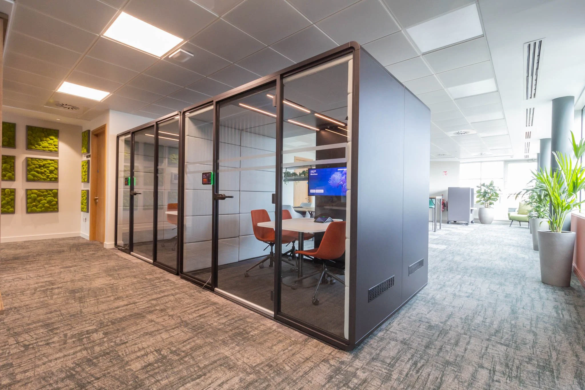 An office space with a glass-enclosed conference room containing a round white table and orange chairs. The room has a large monitor on the wall displaying a blue background. The office has a carpeted floor, potted plants, and large windows letting i