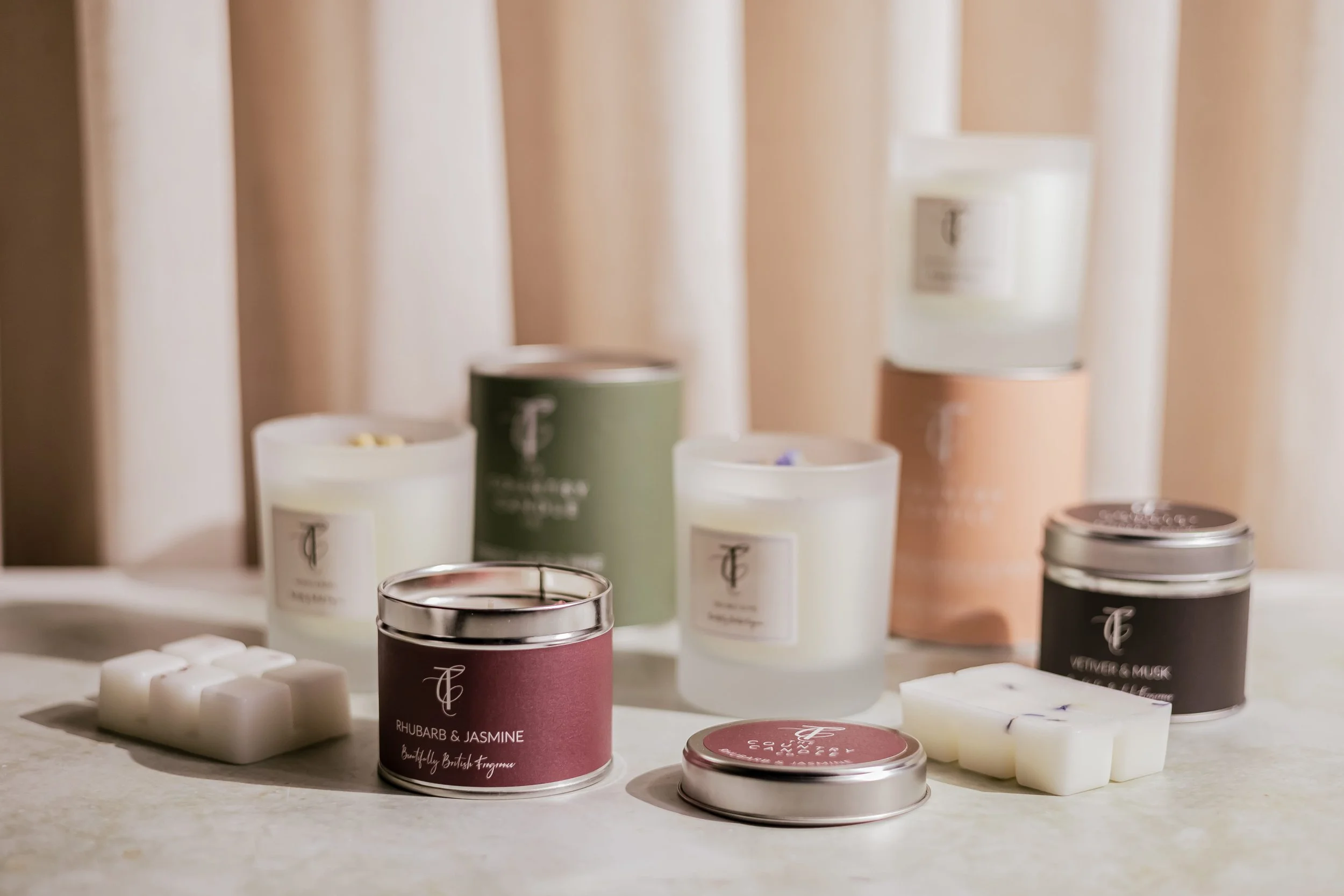 Several scented candles in jars and wax melt cubes on a light-colored surface with beige curtains in the background.