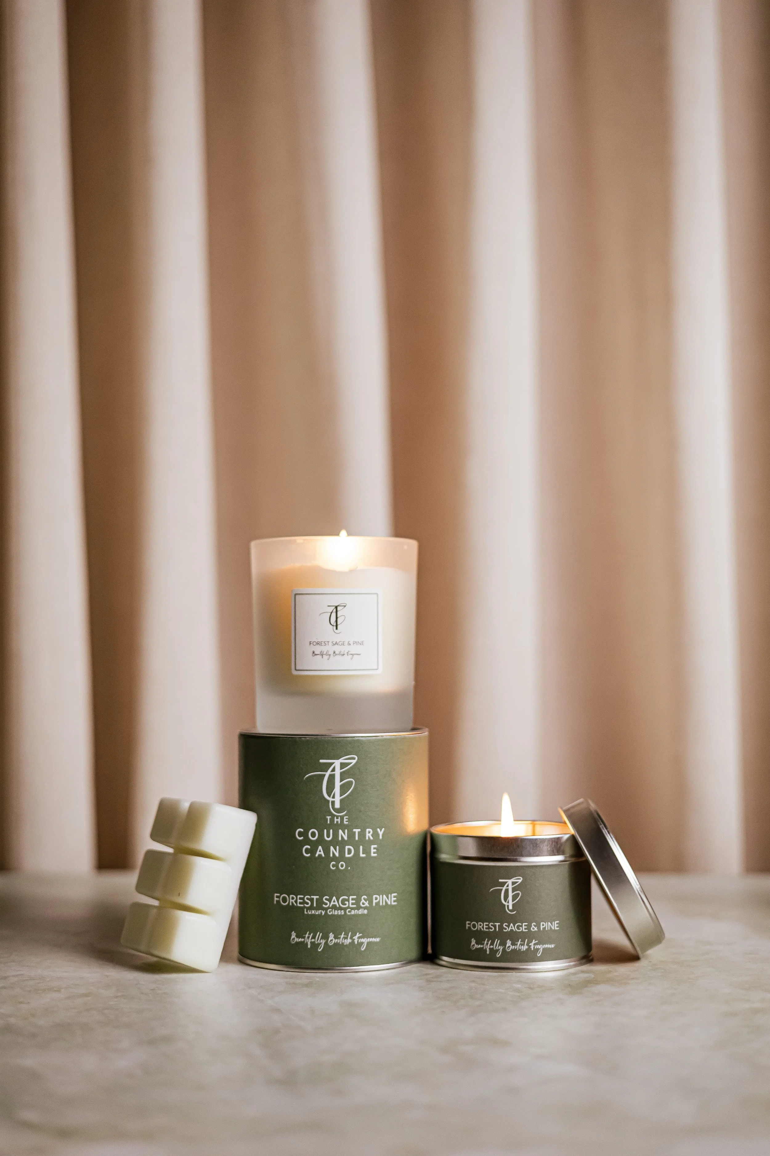 Three candles and three wax melts on a light-colored surface, with a background of beige curtains. The candles are labeled 'Forest Sage & Pine' from The Country Candle Co.