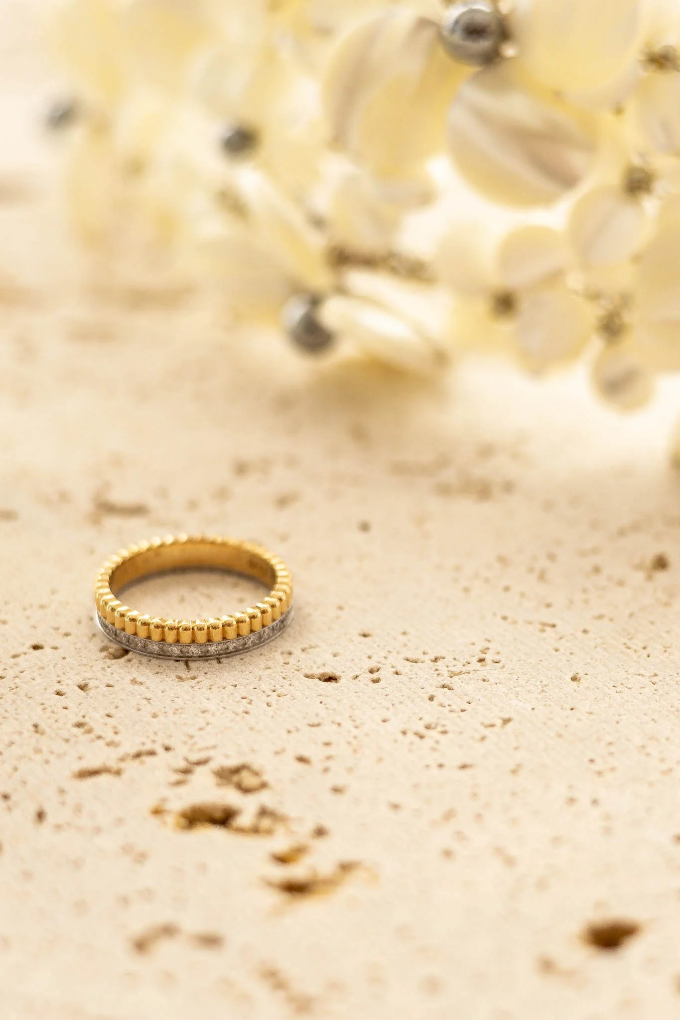 A gold and silver ring on a sandy surface with blurred white flowers in the background.