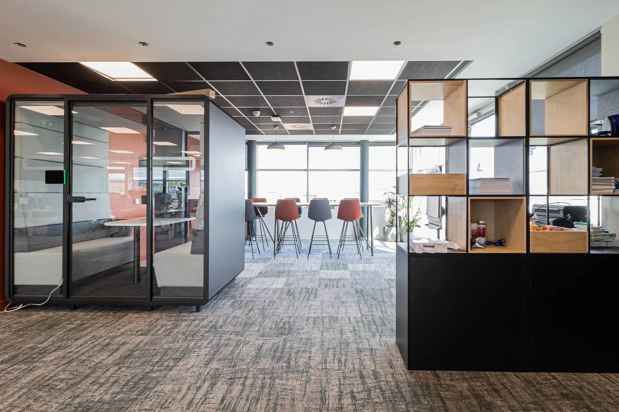 Modern office space with a glass-walled meeting room on the left, a corridor with patterned carpet, and a bar-height table with four chairs near large windows in the background. There are shelving units with books and office supplies on the right sid