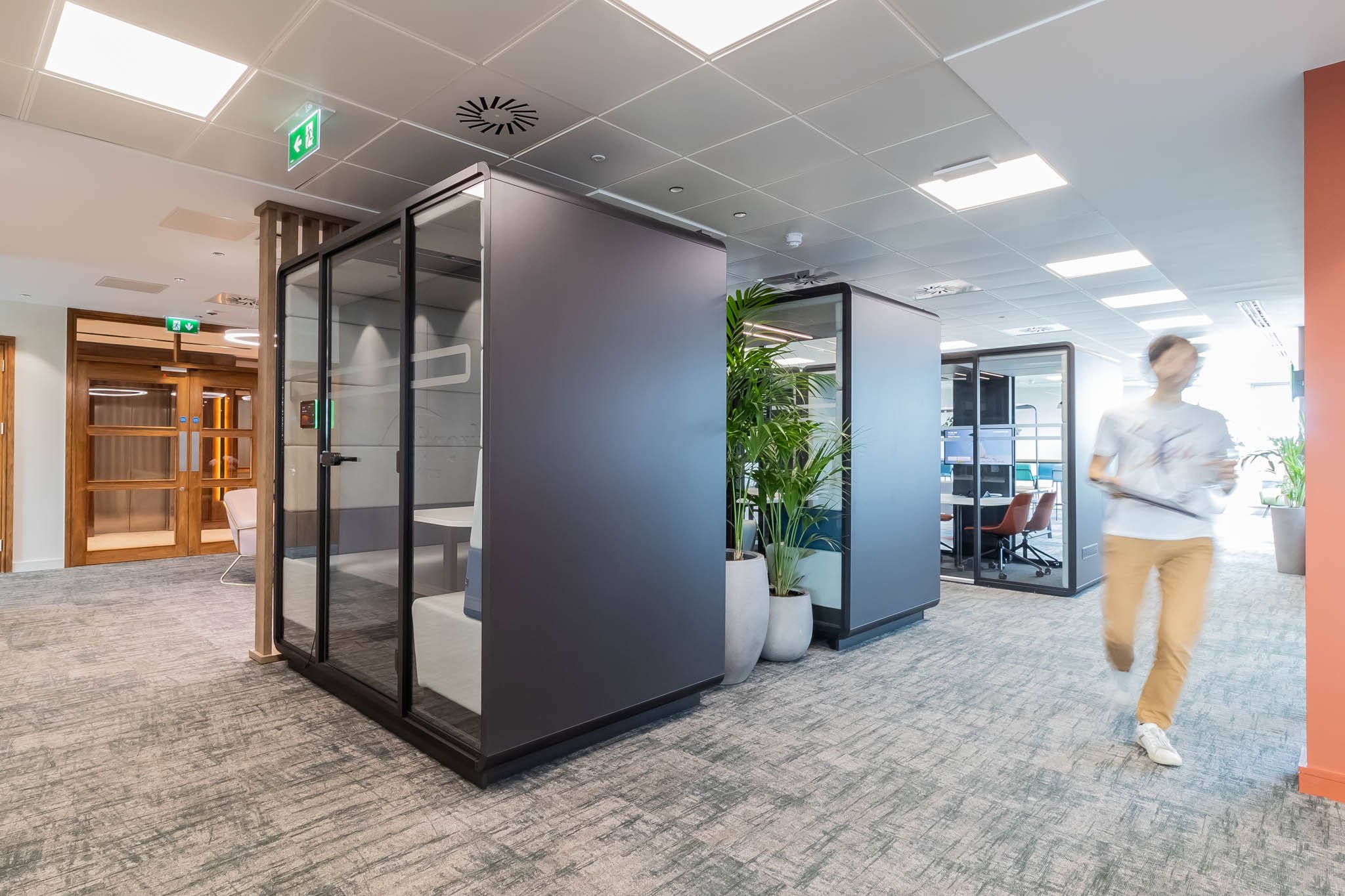Modern office hallway with glass enclosed meeting pods, potted plants, and a person walking by with a tablet.