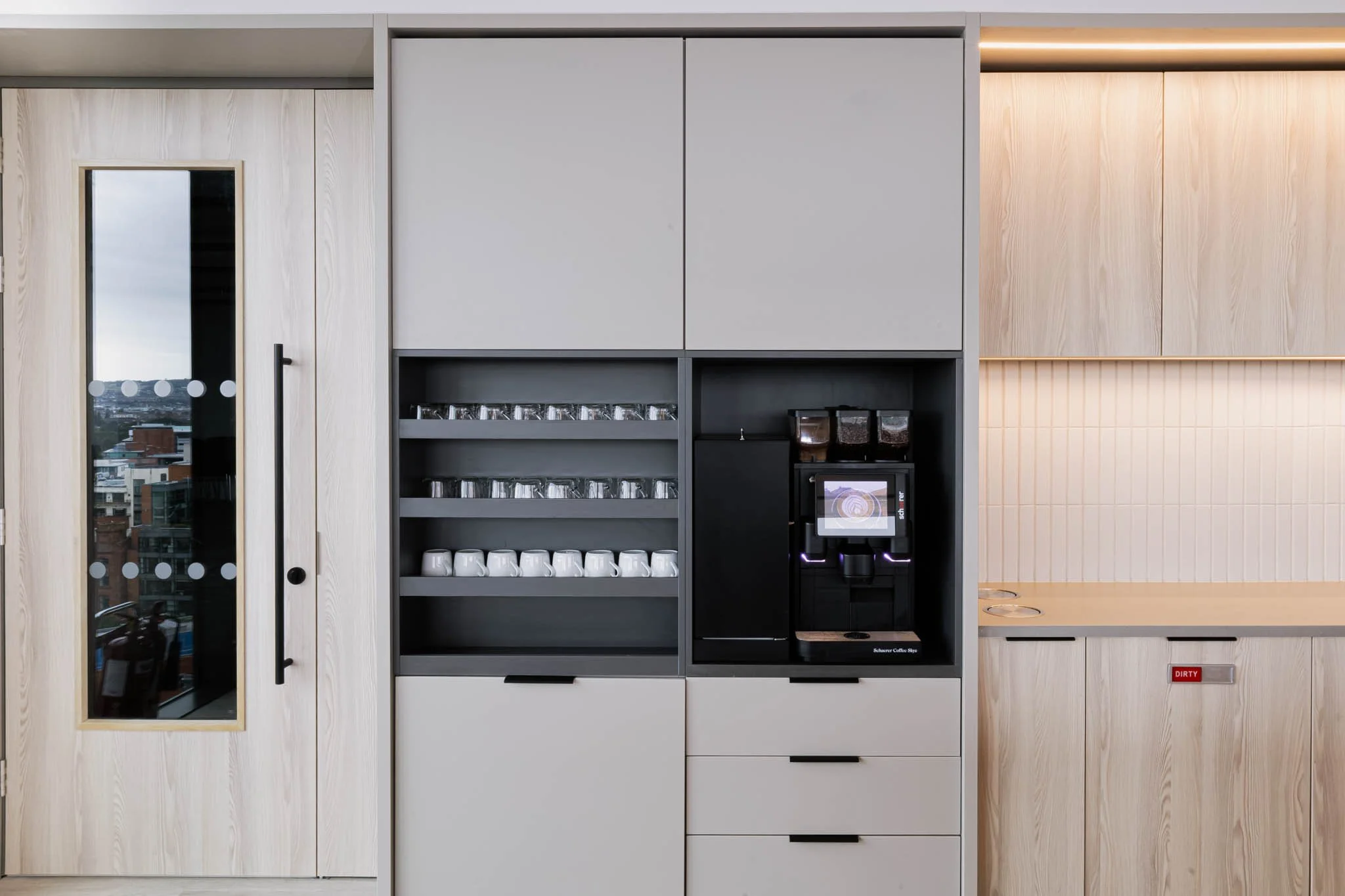 Coffee station with layered shelves of glasses and cups, a coffee machine, and a wooden cabinet with a coffee cup return slot, captured for commercial architecture photography
