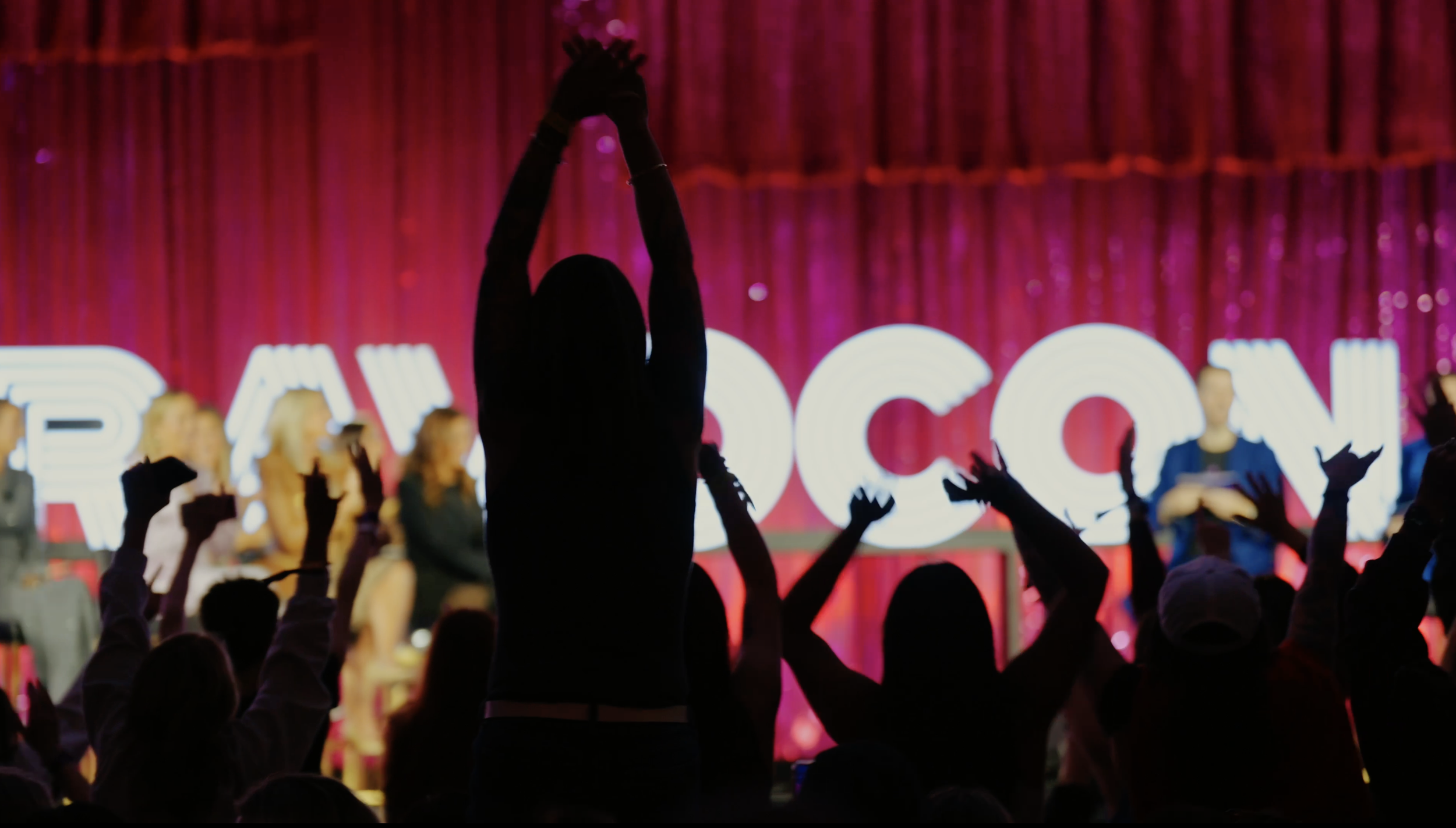 Audience at concert or event with hands raised, silhouetted in front of a stage with large illuminated letters and a red curtain background.