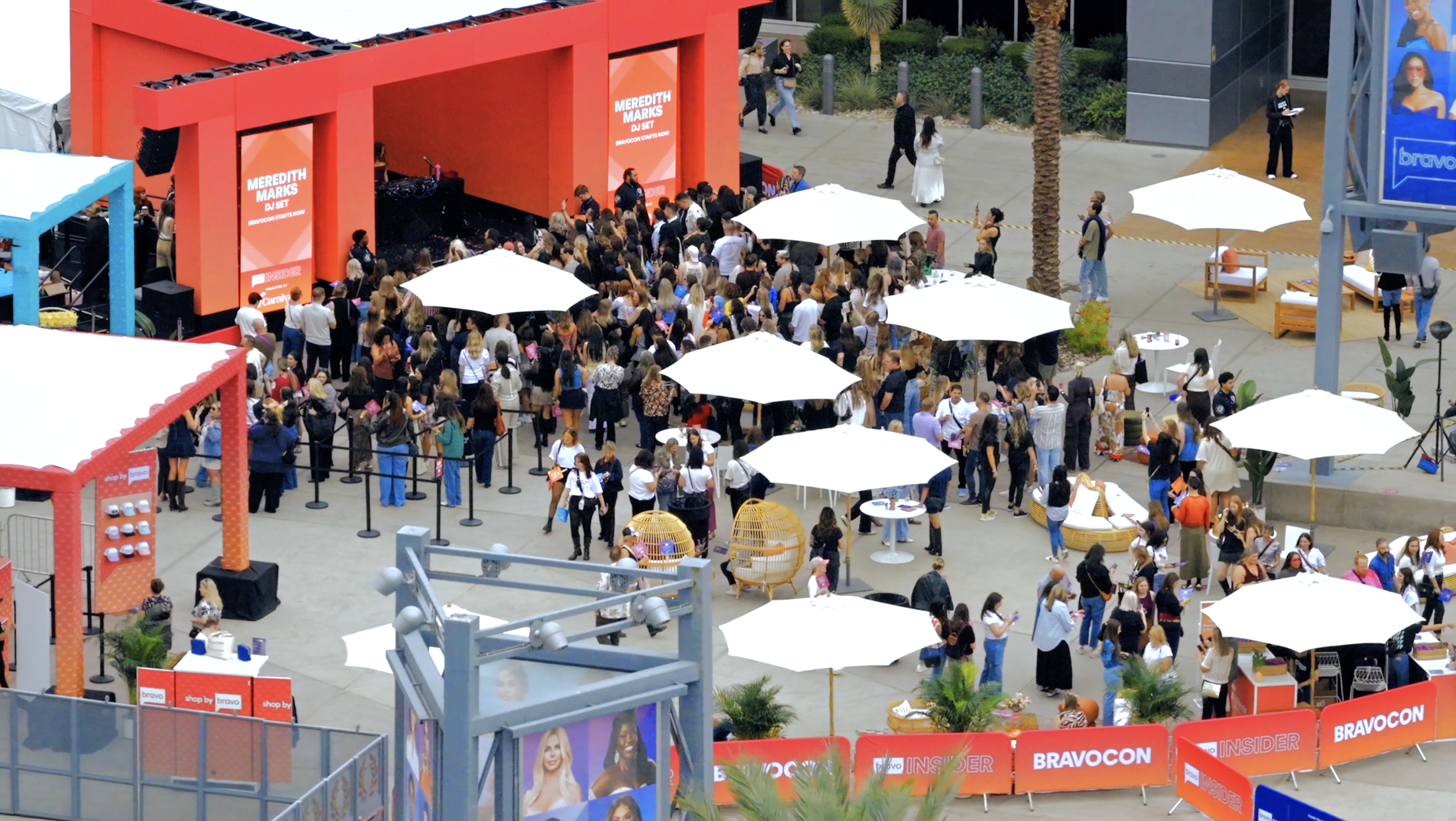 An outdoor event with a large crowd gathered in front of a stage with red panels, showing a DJ set by Meredith Marks. There are white umbrellas providing shade, seating areas, and various displays, with some people taking photos and others waiting in