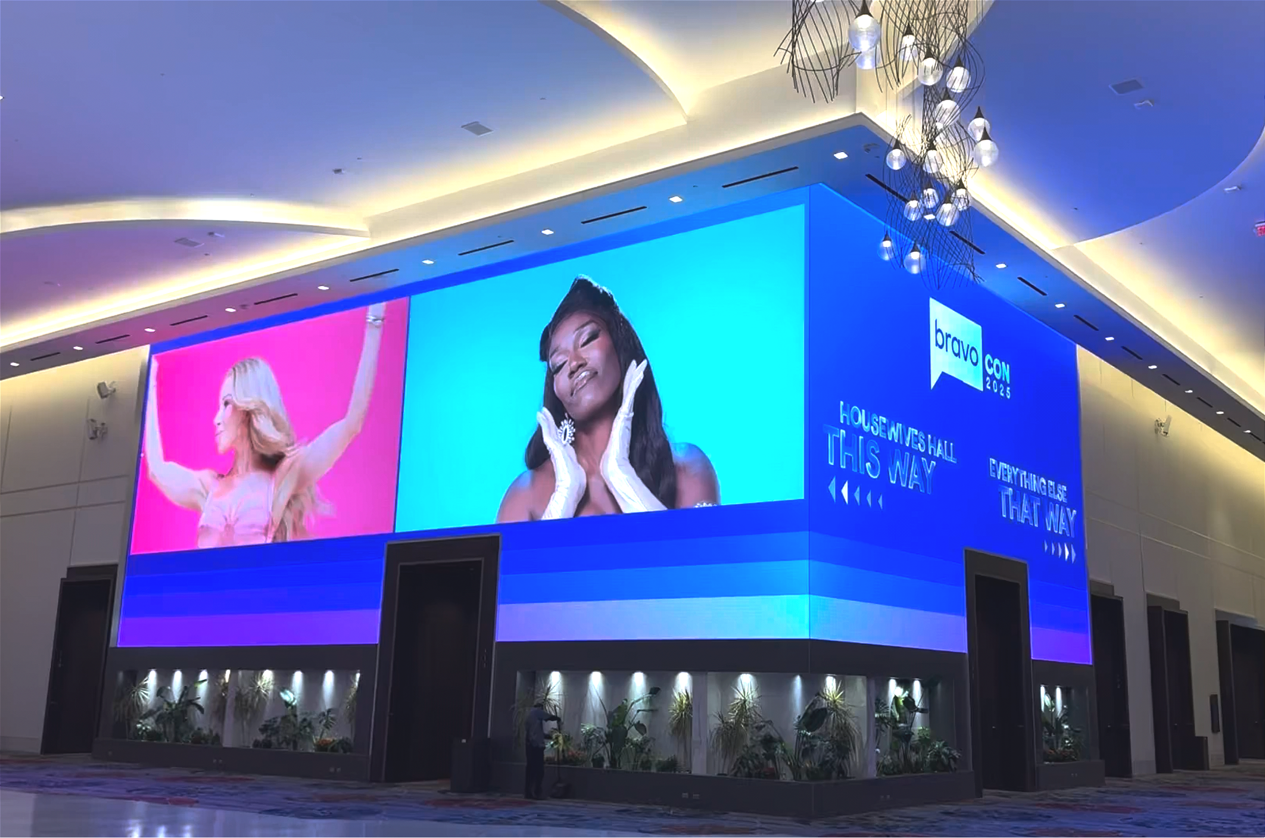 Large digital screen in a conference hall displaying images of three women, one with blond hair and pink background, one with black hair and blue background. Text on the screen reads 'Bravo Con 2025,' 'Housewives Hall,' 'This Way,' and 'Everything El