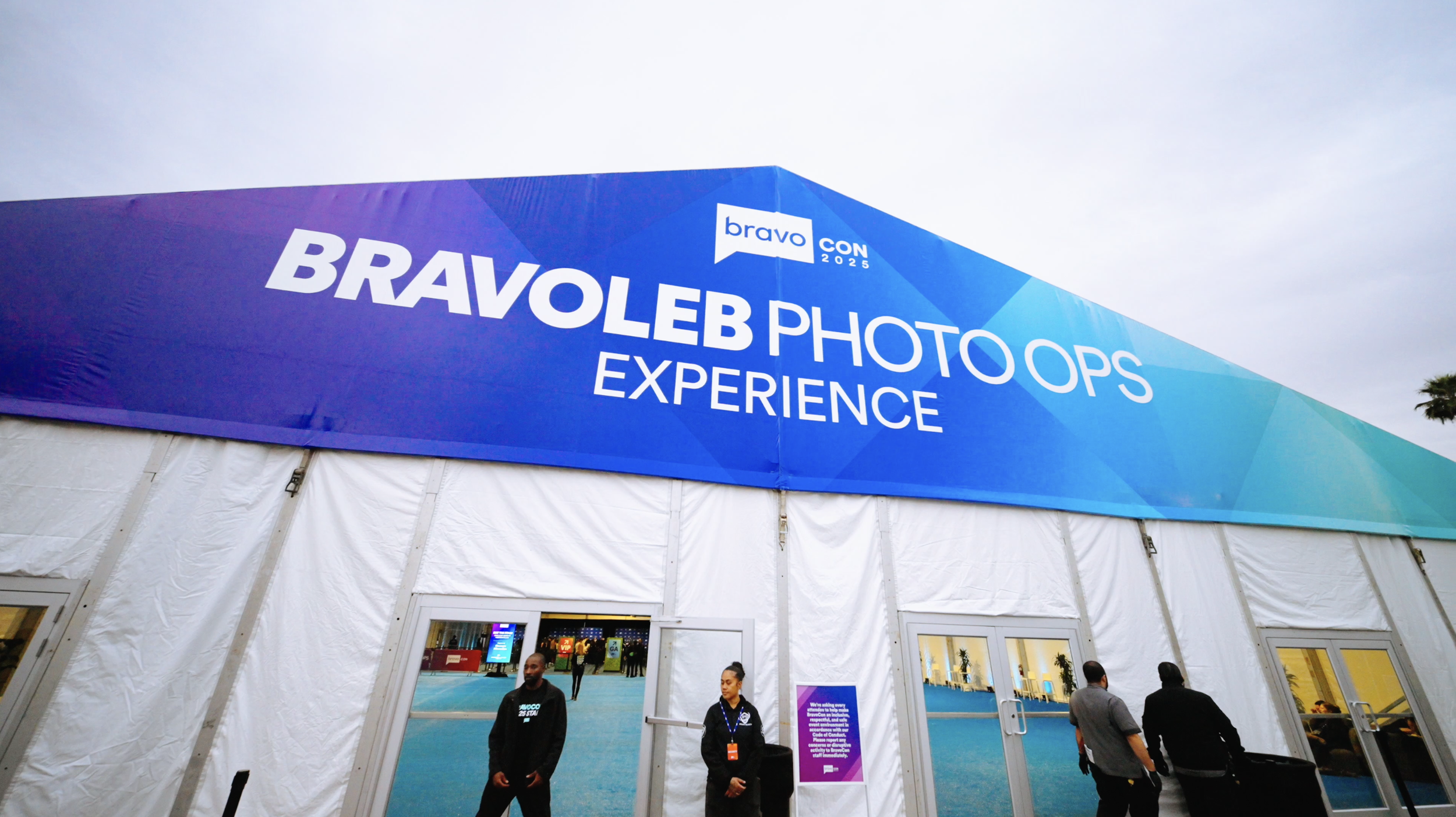 Large blue banner at BravoCon 2025 promoting Bravo Photography Experience with people walking in front of it.
