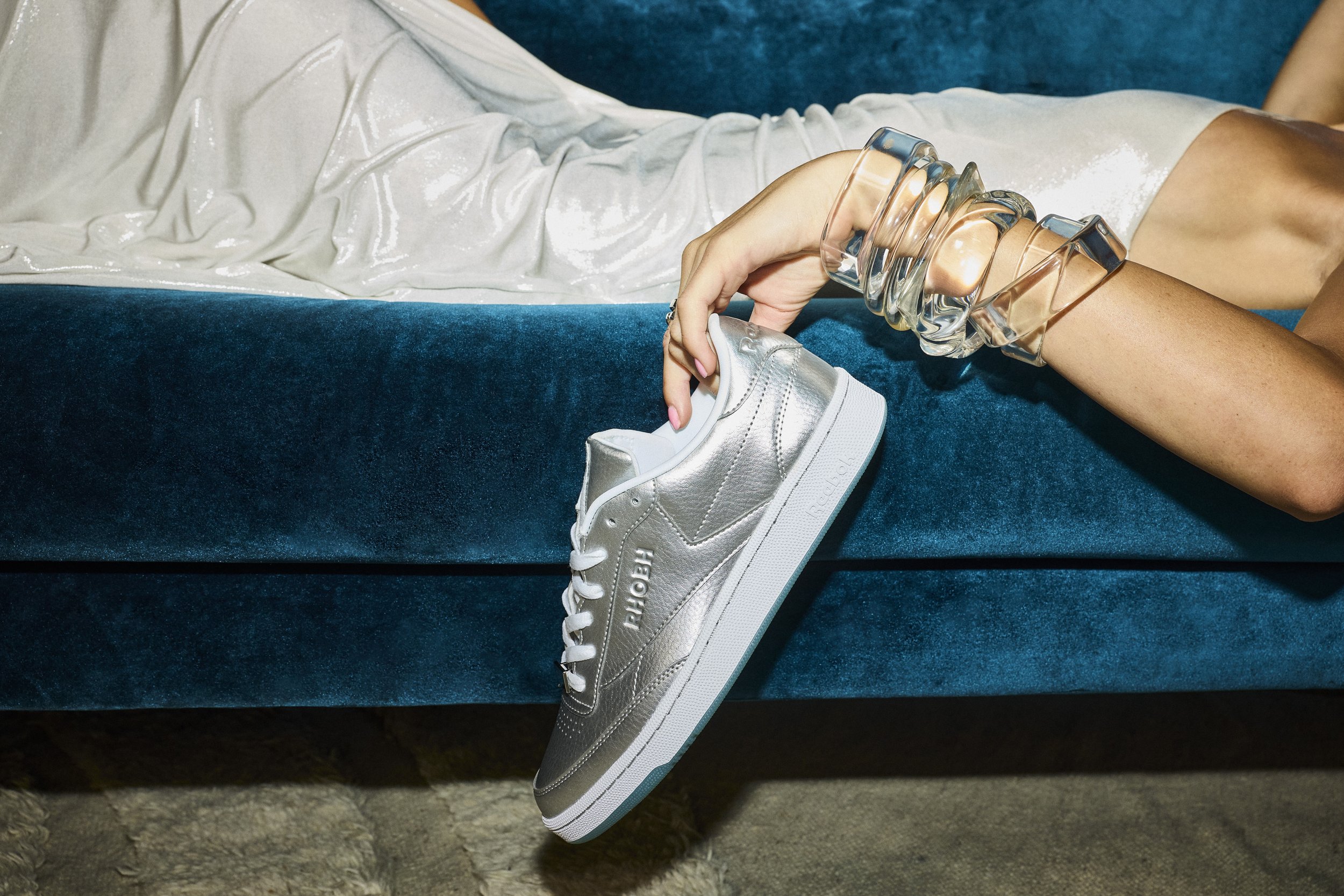 A person sitting on a blue velvet sofa, holding a silver sneaker with white laces, while wearing multiple chunky transparent bracelets on their arm.