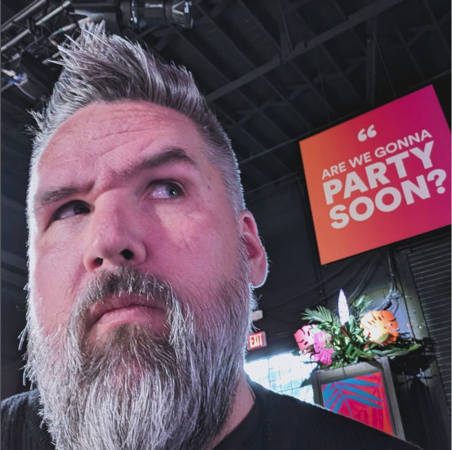 Close-up of a man with gray beard and spiked hairstyle looking puzzled or suspicious, in a party or event venue with a colorful sign in the background that says, "Are we gonna party soon?" decorated with tropical flowers and leaves.