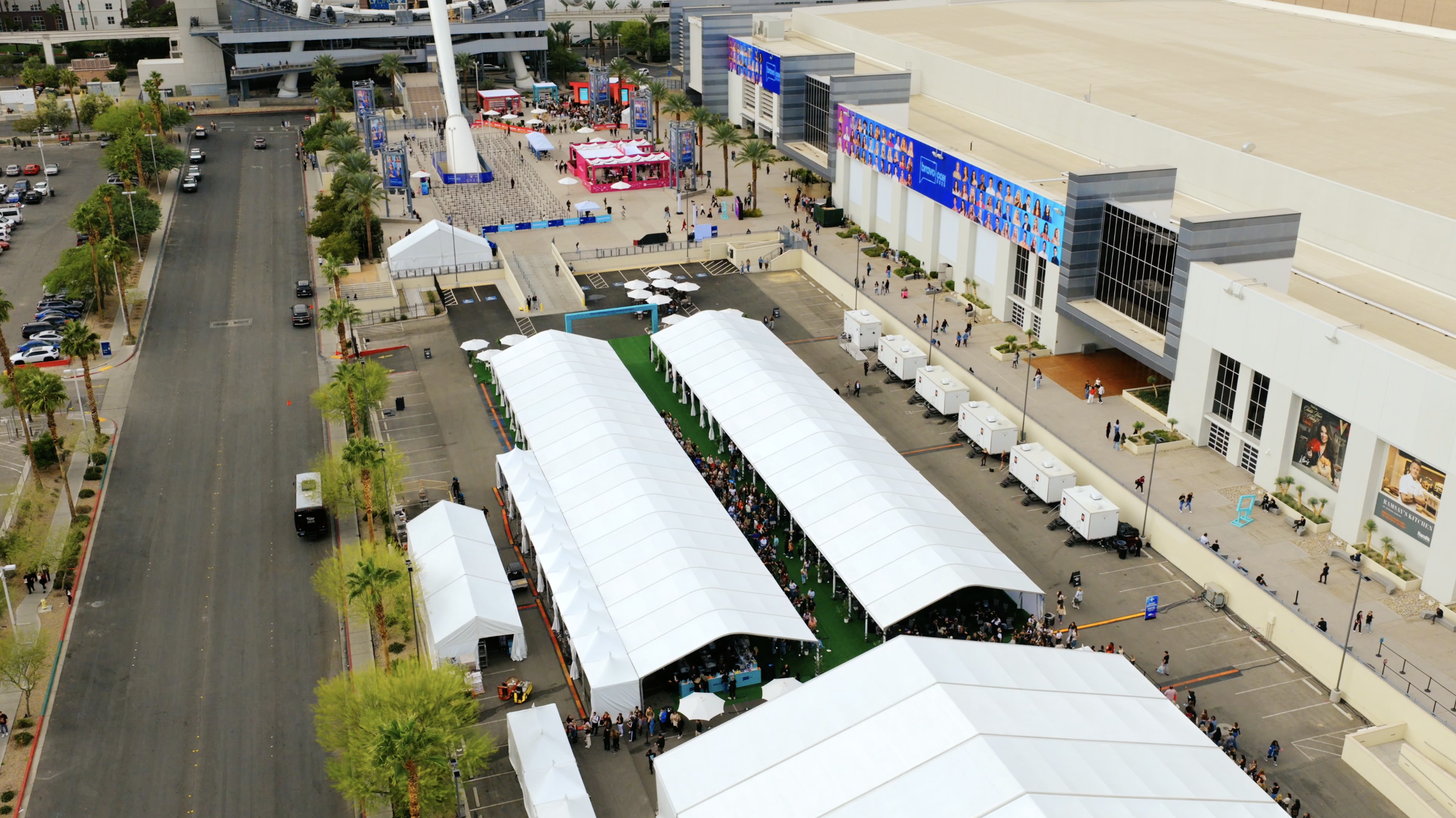 Aerial view of a large convention center with white tents and a crowd of people, event booths, and outdoor screens on the building facade.