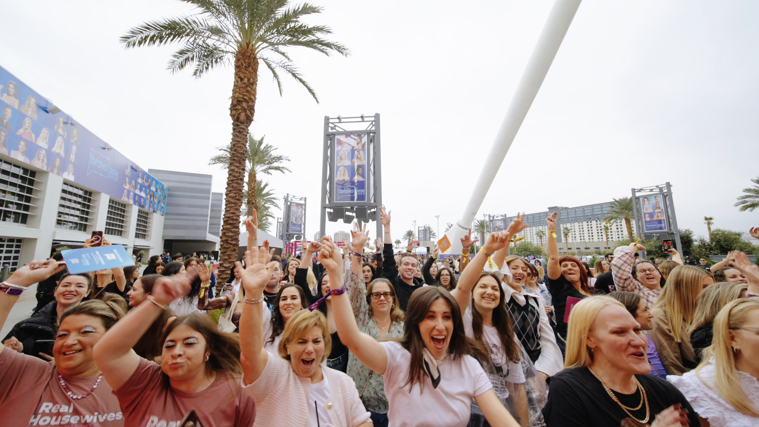 A crowd of people outdoors, smiling and cheering at a daytime event with palm trees and large screens displaying a TV show.
