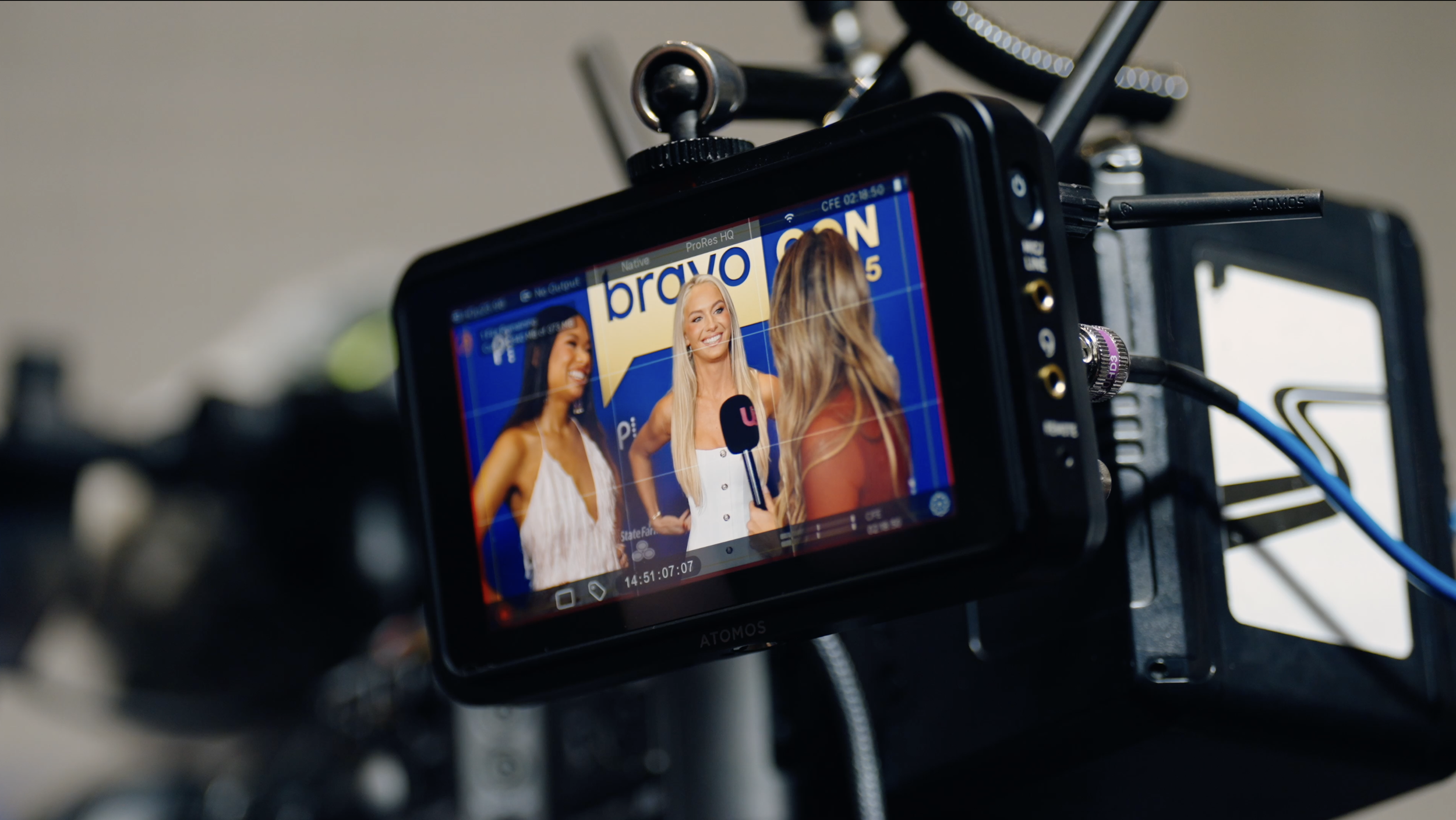 Three women are being interviewed on camera with a backdrop that says 'bravo' and 'on'. One woman with long blonde hair holds a microphone and is smiling, while the other two women, one with dark hair and the other with light brown hair, are smiling 