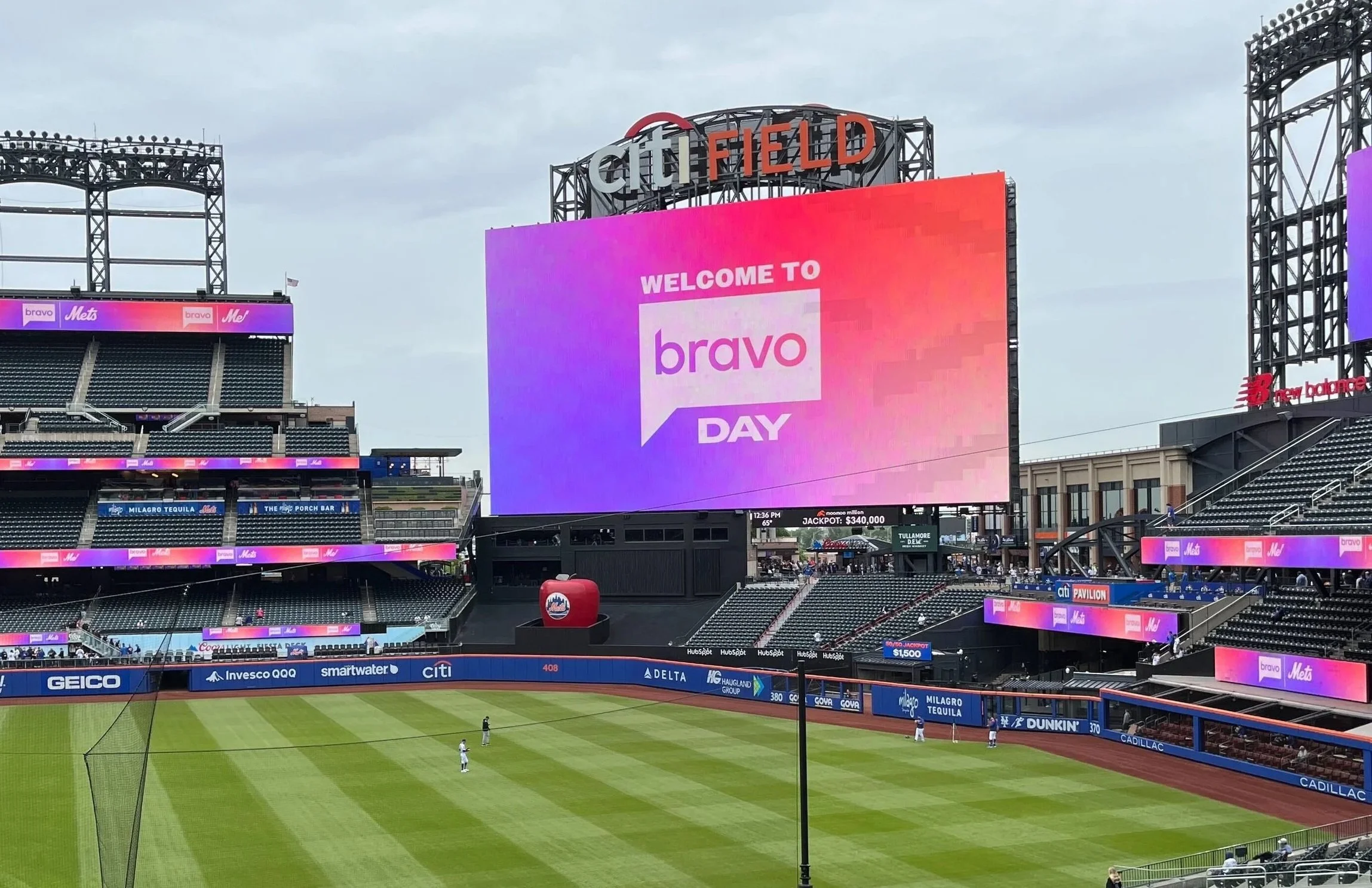 A baseball stadium with a large digital scoreboard displaying a pink and purple welcome message for Bravo Day, with players on the field.