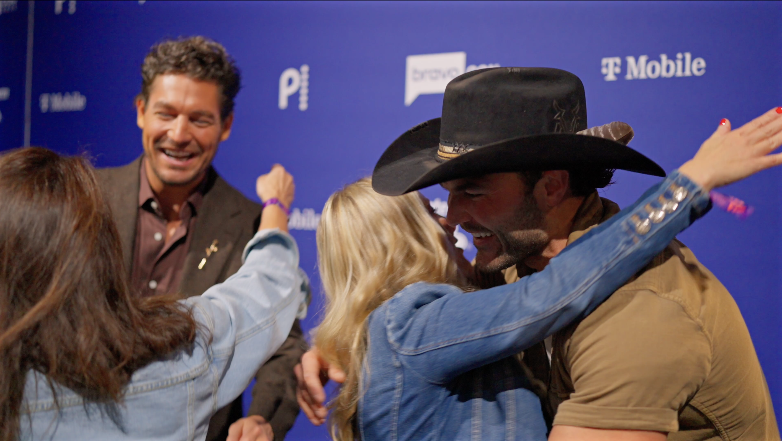 A group of people hugging and smiling at an event with a blue backdrop displaying logos.
