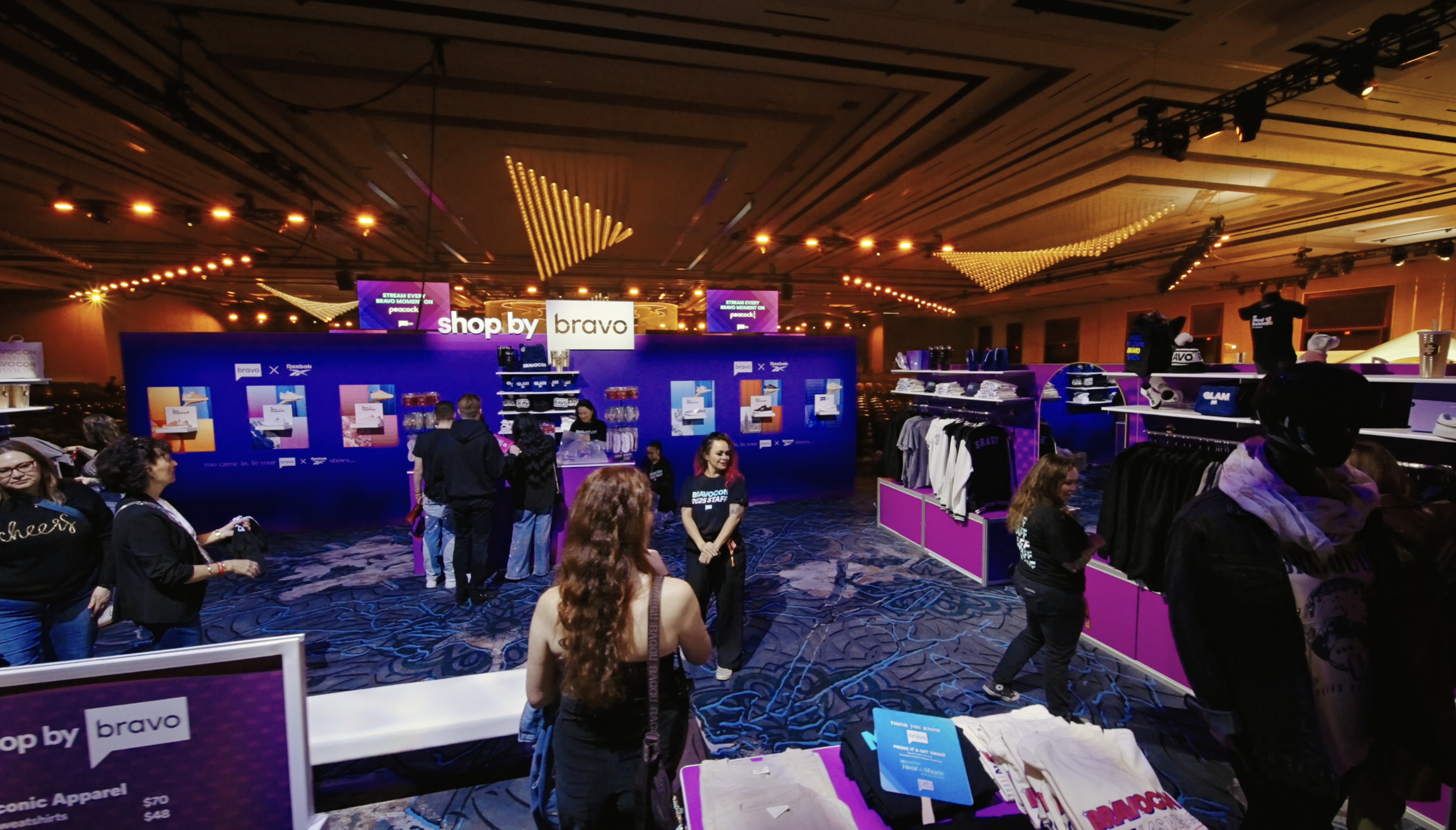 People shopping at the Bravo x Reebok apparel booth in a large, well-lit conference hall with purple and yellow accents.