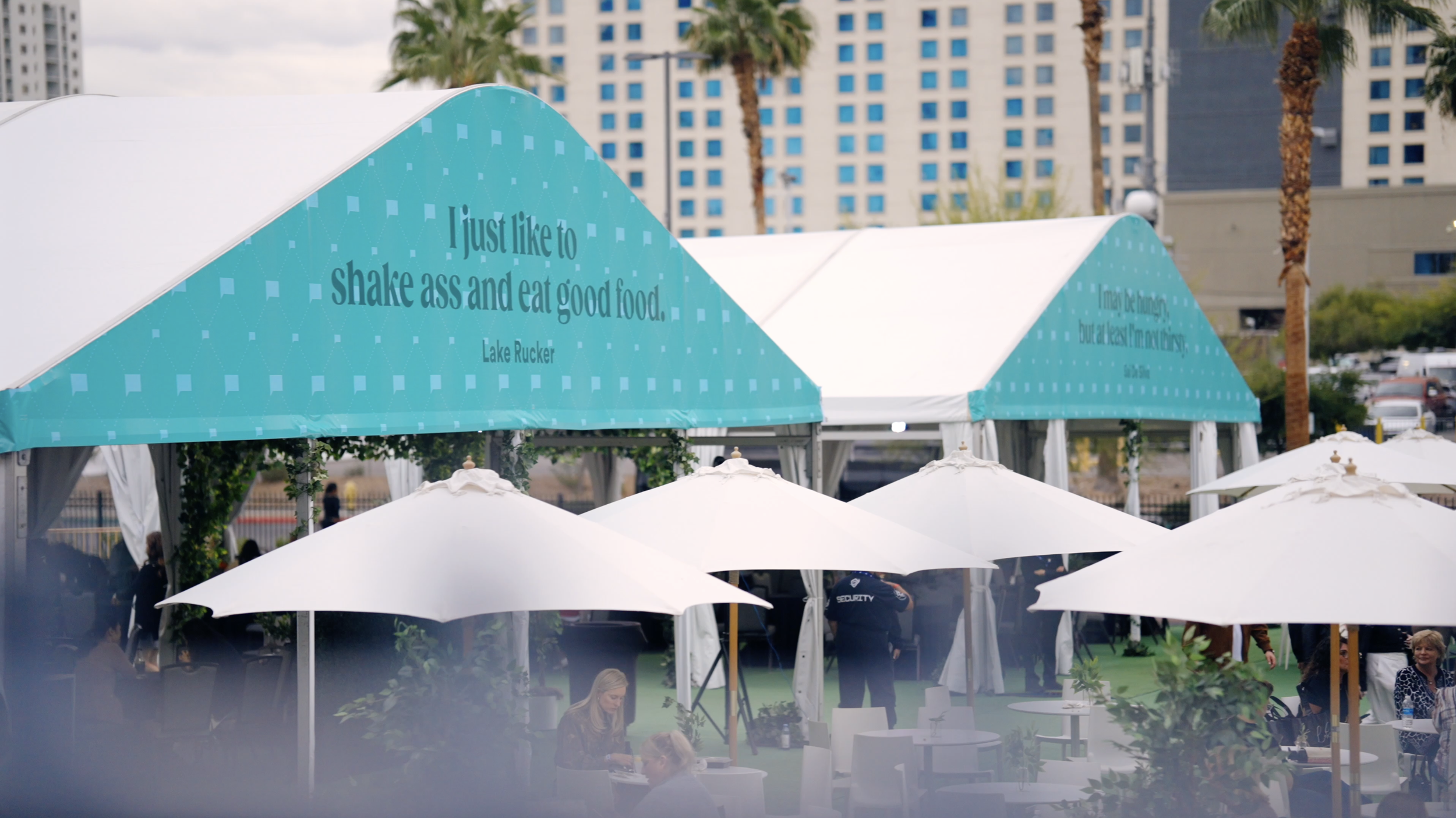 Outdoor event with white umbrellas, tables, and people. There are tents with teal banners displaying humorous quotes about food and fitness. Background shows palm trees and tall buildings.