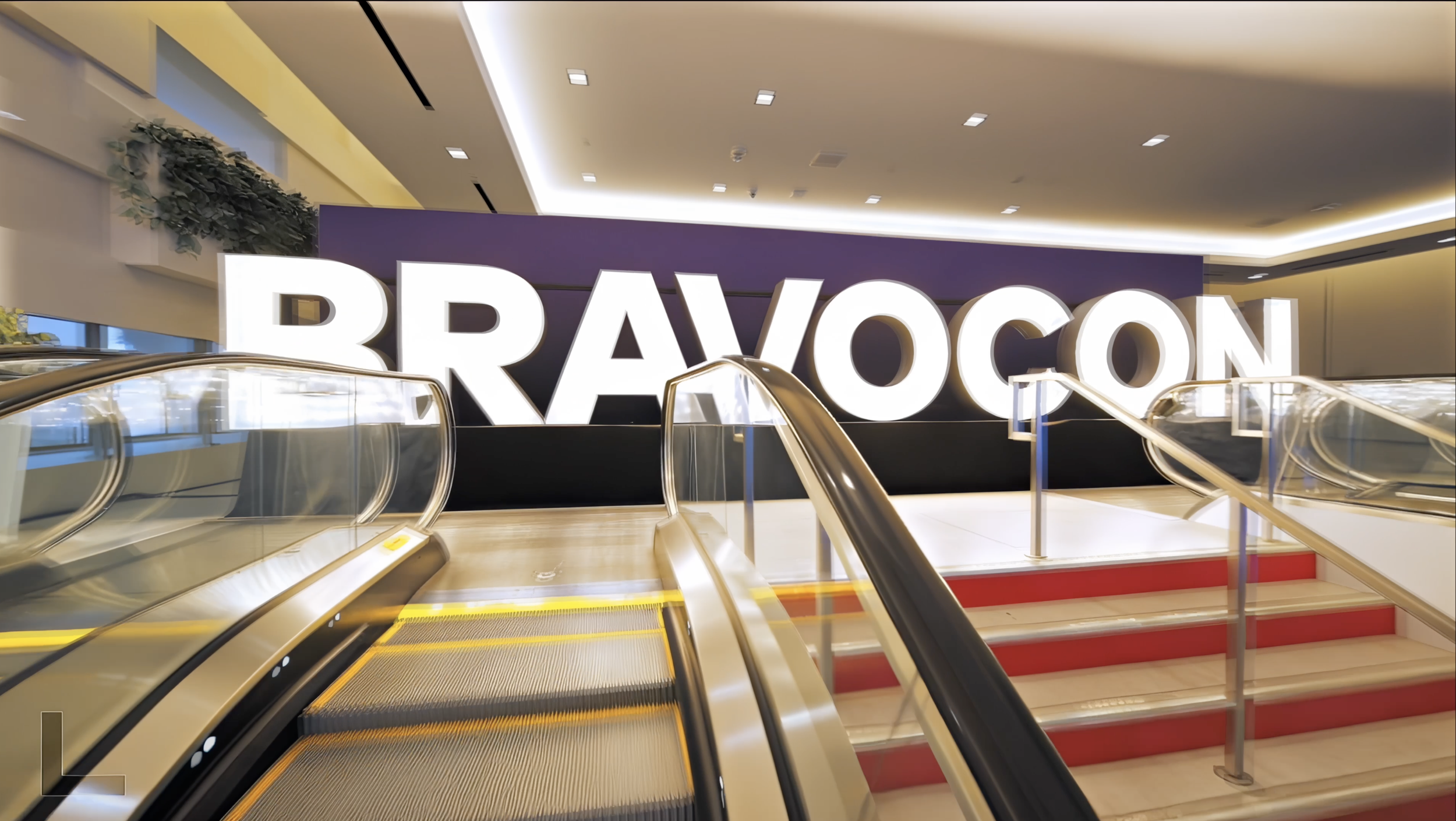 Interior of a modern building with a large illuminated sign spelling 'BRAVOCON' behind an escalator and staircase.