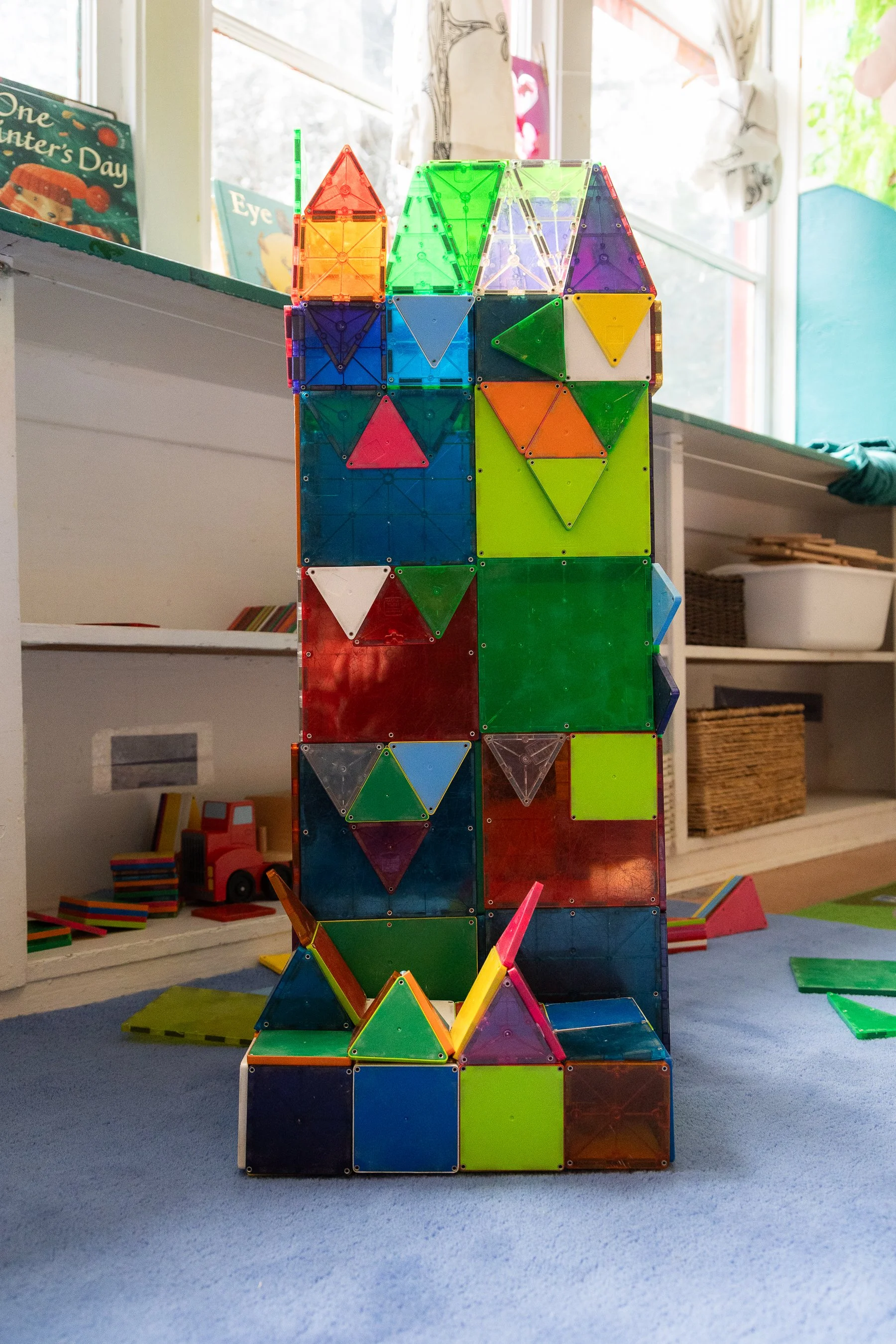 Colorful magnetic building blocks arranged in a tower on a blue carpeted floor in a playroom, with books and toys in the background.