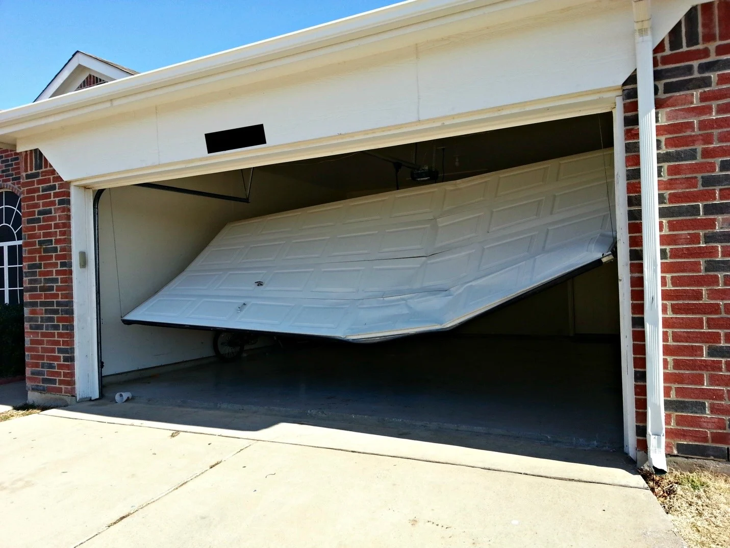 A garage door in the process of opening or closing, revealing a partially raised white sectional garage door. The garage is attached to a brick house, and the driveway extends in front of it. The sky is clear and blue.