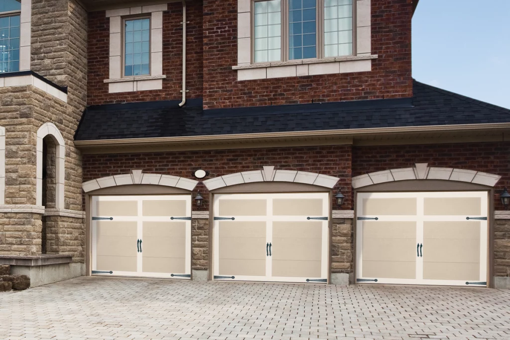 Three beige garage doors with black handles and hinges in front of a brick house with stone accents and upper windows.