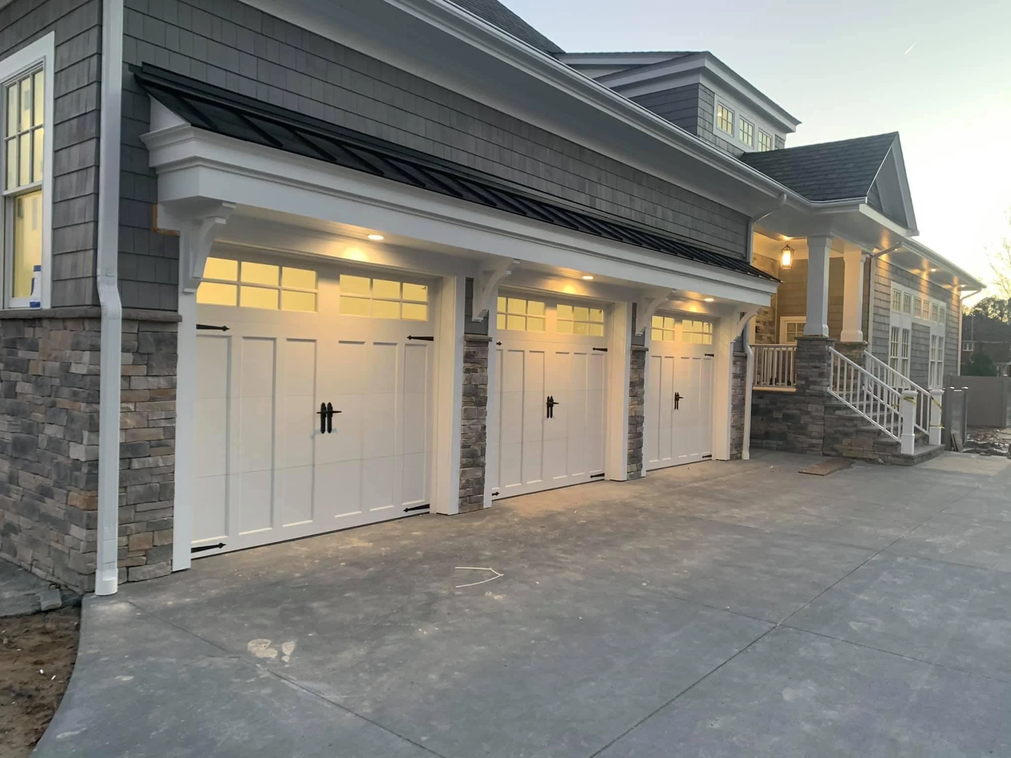 A three-car garage with white doors, stone accents, and a porch with stairs leading to a house in the background, illuminated by outdoor lights at dusk.