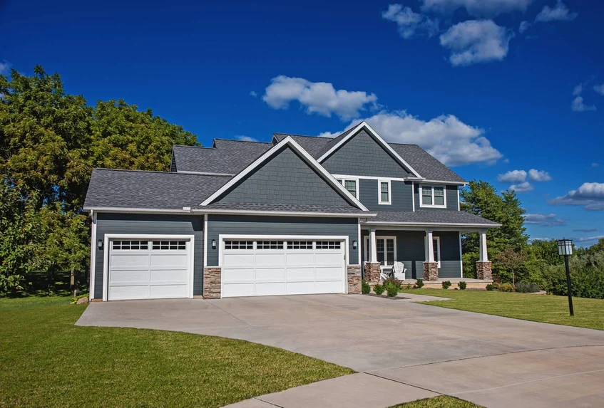 A large blue two-story house with three garage doors, a driveway, a front porch with white chairs, green lawn, trees, and a blue sky with clouds.