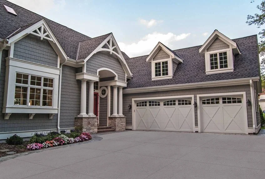 Front view of a large gray house with a three-car garage, dormer windows, a covered front porch with columns, and a flower bed with colorful plants.