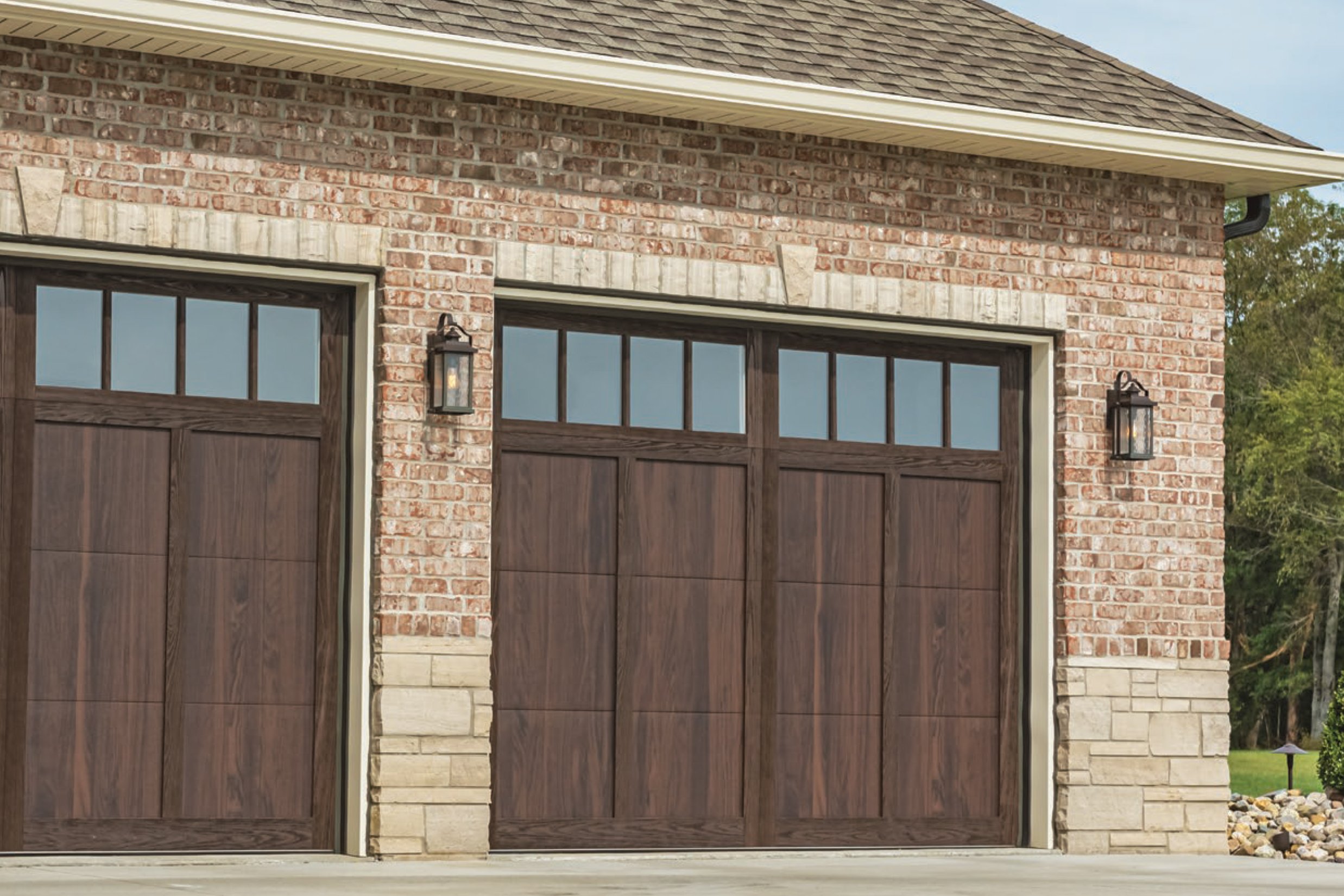 A garage with two wooden doors and brick exterior, featuring black lantern-style lights on each side, and a stone accent at the bottom of the brick wall.